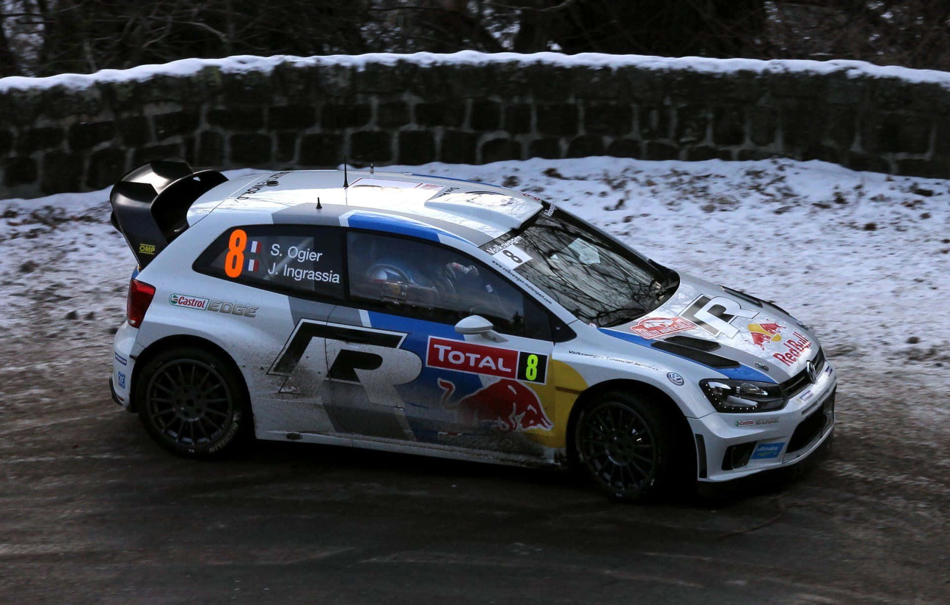 Ice Racing 桌布 壁紙 — 一輛帶有 Red Bull 和 Total 品牌標誌的白色拉力賽車沿著被石牆環繞的積雪路邊賽道疾馳。