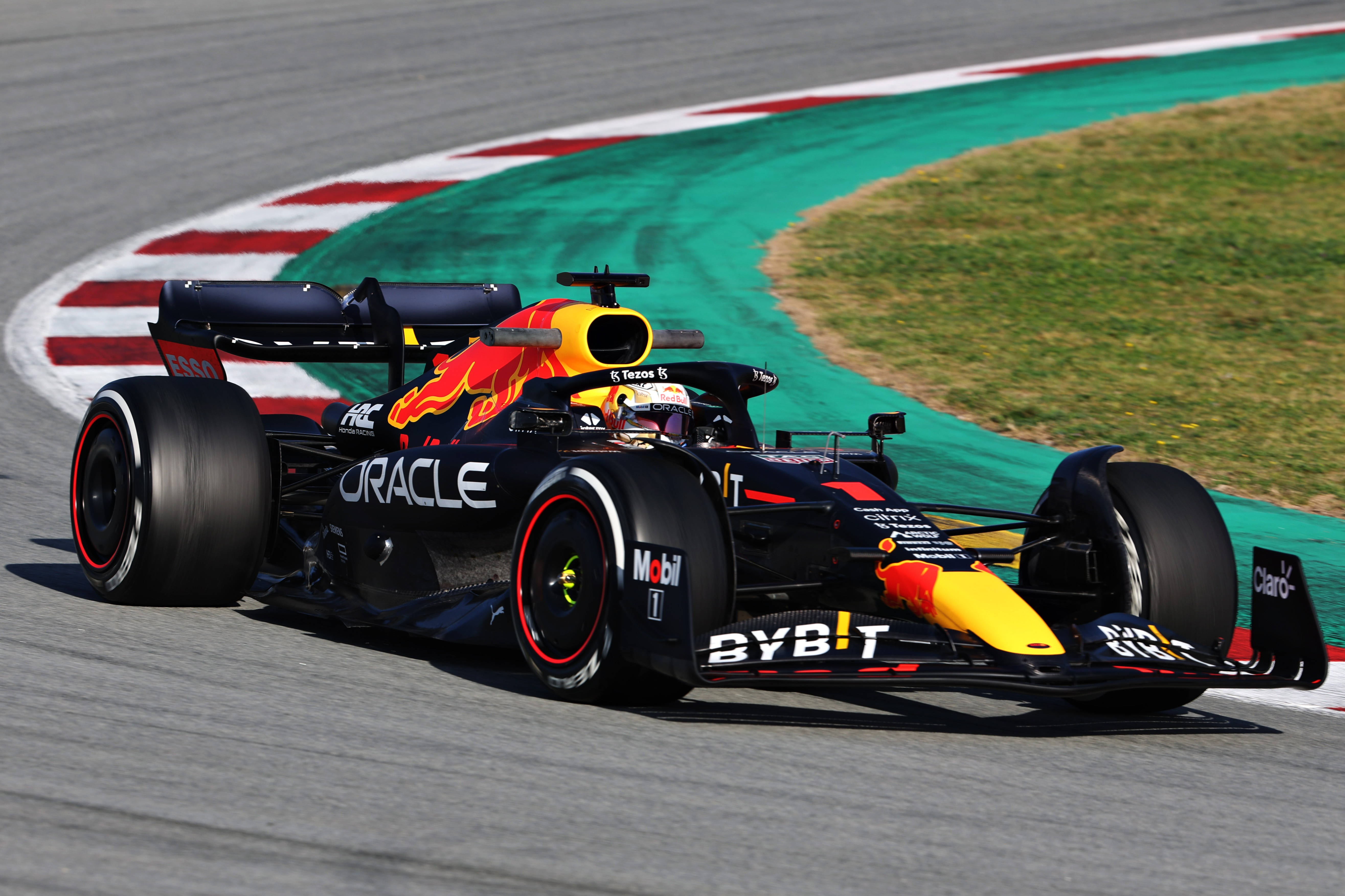 Verstappen 桌布 壁紙 — 一輛帶有Oracle和BYBIT品牌標誌的Red Bull一級方程式賽車，在陽光明媚的賽道上疾馳過一個急彎，賽道兩旁是綠色草地和紅白相間的賽道邊緣。