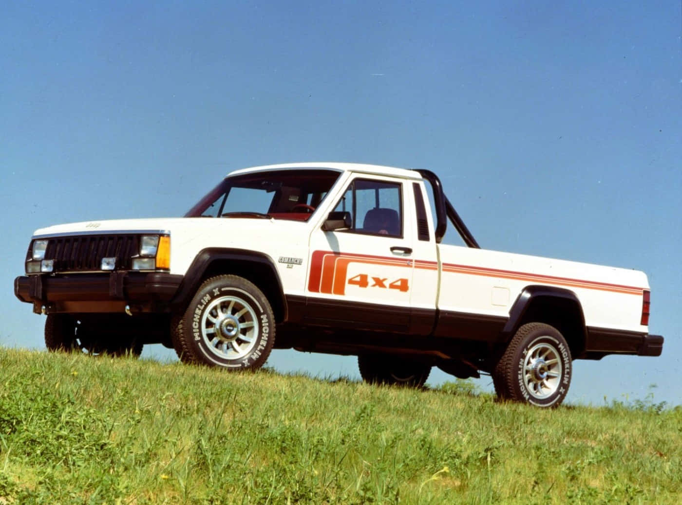 Jeep Comanche 桌布 壁紙 — 一輛帶有橙色條紋的白色 4x4 皮卡車停在晴朗藍天下長滿草的山丘上。