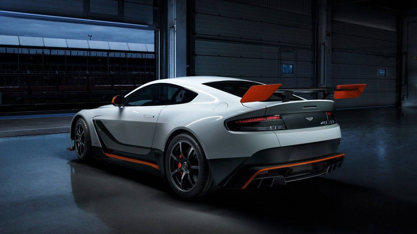HD 桌布 壁紙 — 一輛帶有橙色點綴的時尚白色 Aston Martin Vantage GT12 在車庫燈光下閃閃發光，展現其空氣動力學設計和大型尾翼。