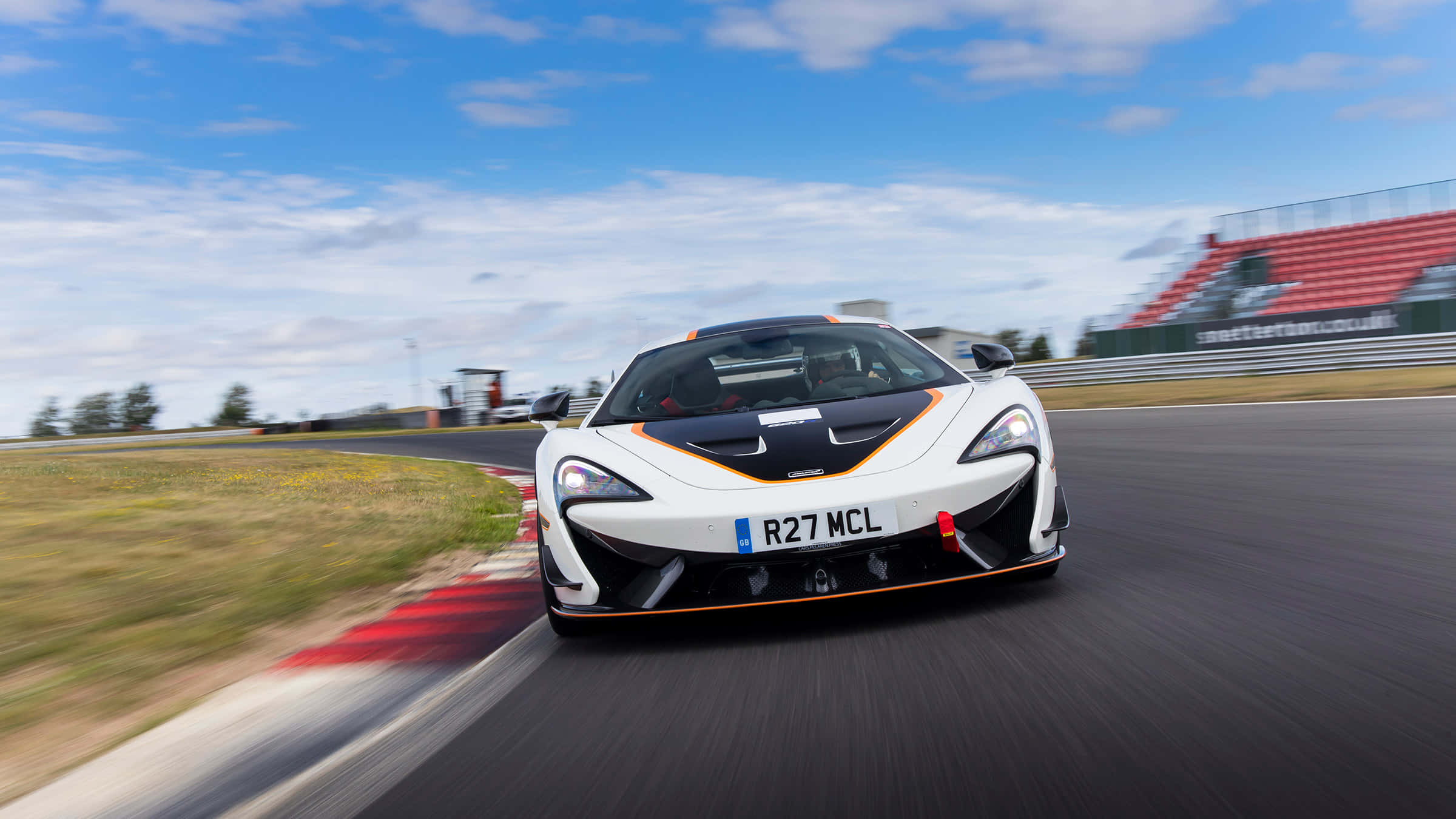 Mclaren 620r 桌布 壁紙 — 一輛帶有橙色點綴的白色McLaren跑車在明亮的藍天下，以動態模糊效果在賽道上飛馳。