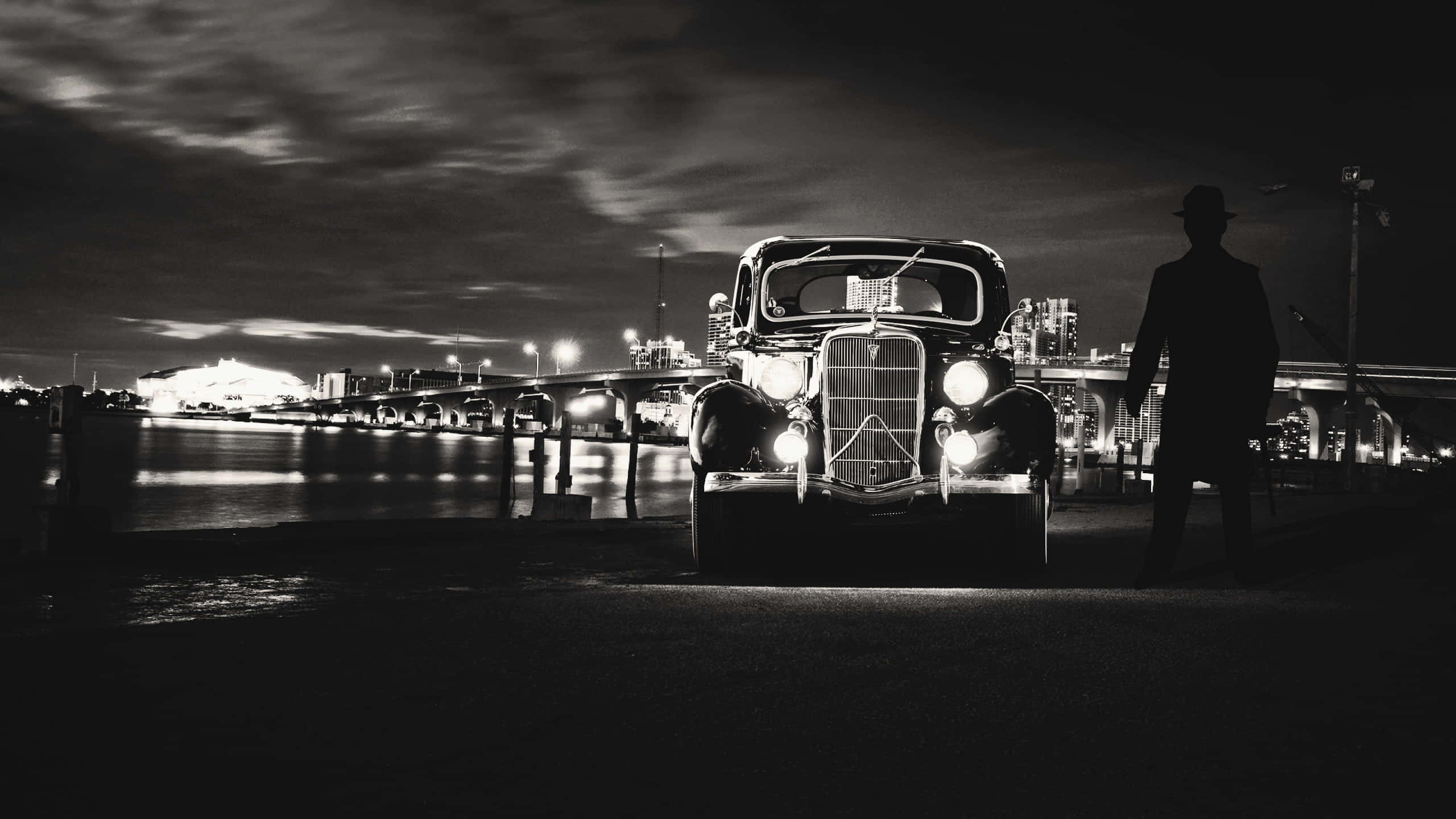 Vintage Black And White 桌布 壁紙 — 一輛帶有發光車頭燈的復古汽車在夜晚停在海濱大橋附近，旁邊有一個剪影男子，天空多雲。