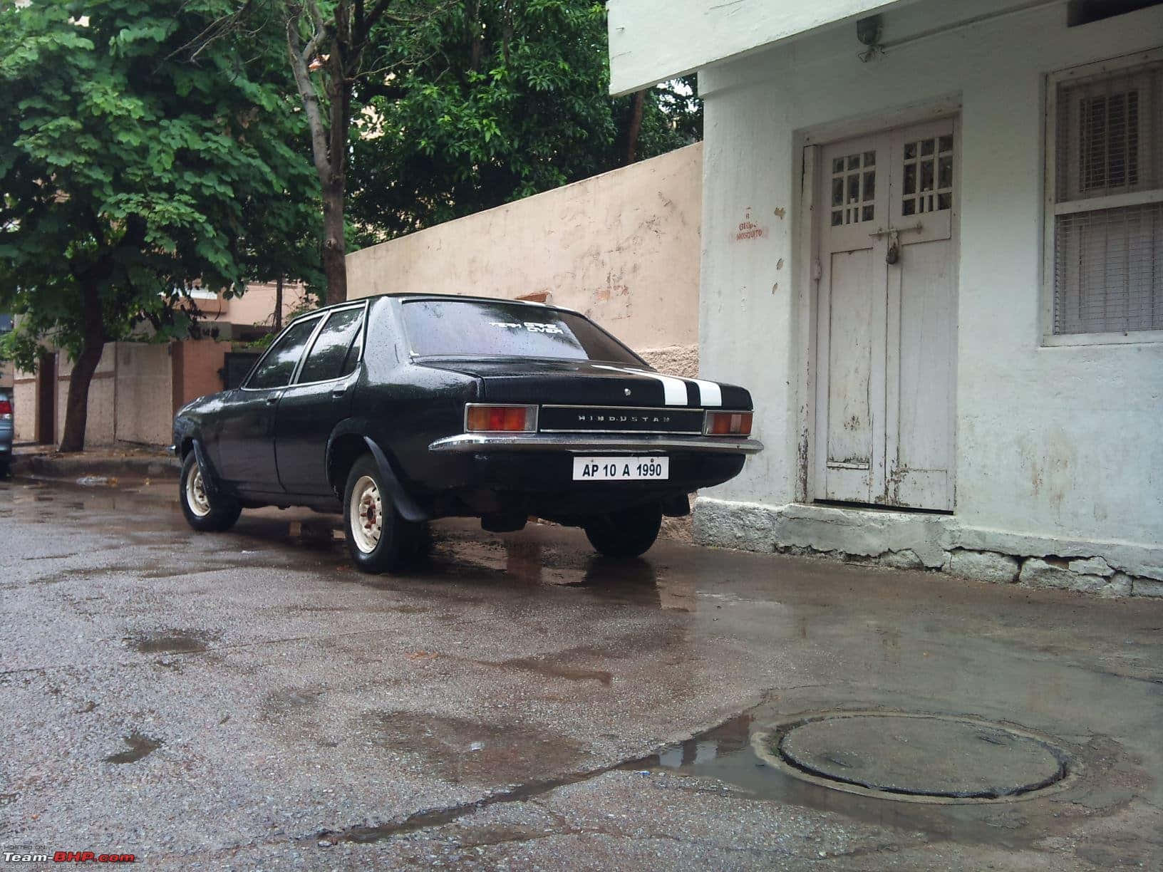 Hindustan Motors 桌布 壁紙 — 一輛帶有白色條紋的黑色復古Hindustan汽車停在潮濕的瀝青街道上，旁邊是一座風化的白色建築。