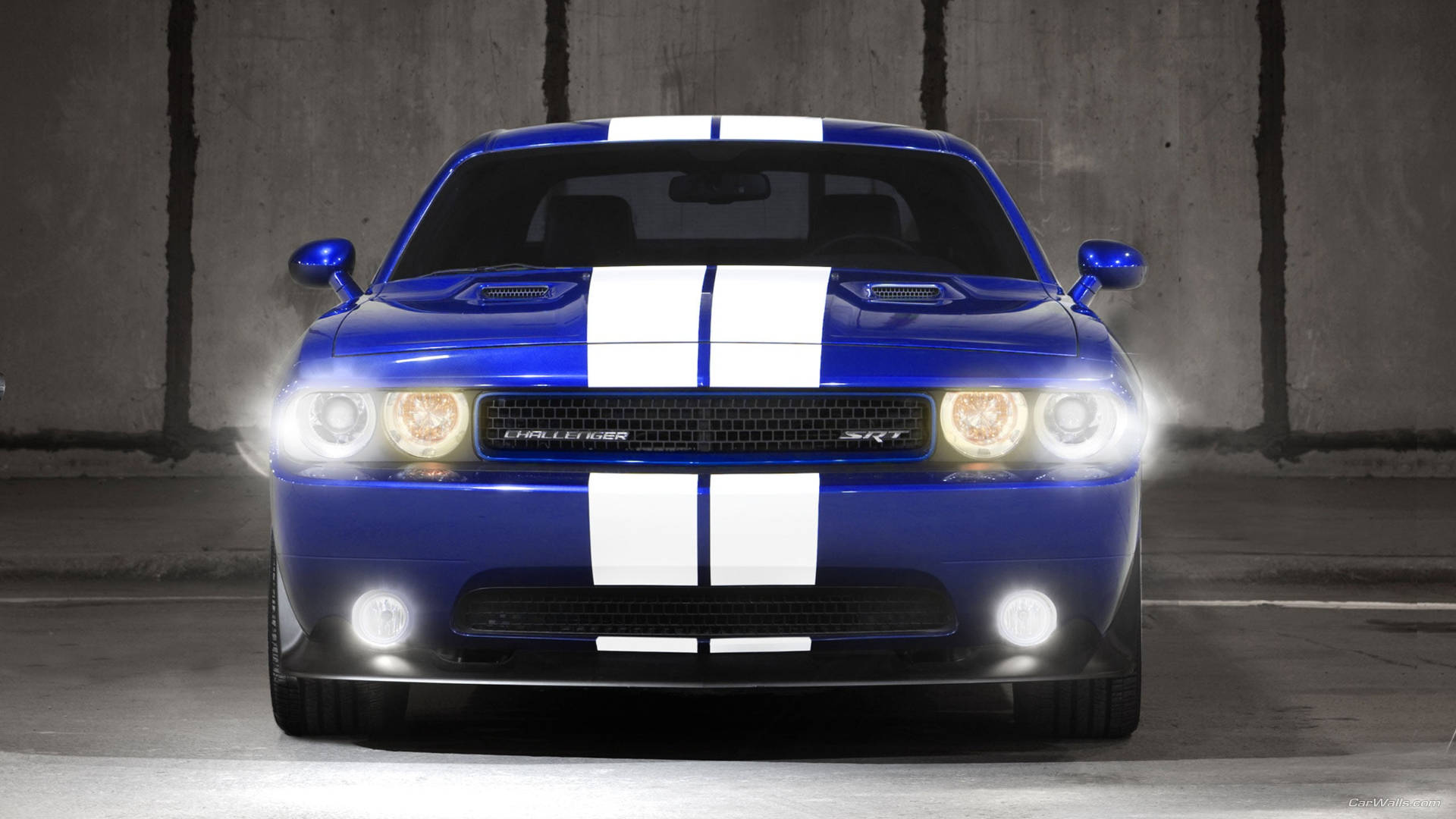 Dodge 桌布 壁紙 — 一輛帶有白色賽車條紋的充滿活力的藍色道奇挑戰者，在昏暗的混凝土車庫中，發光的車頭燈直視前方。