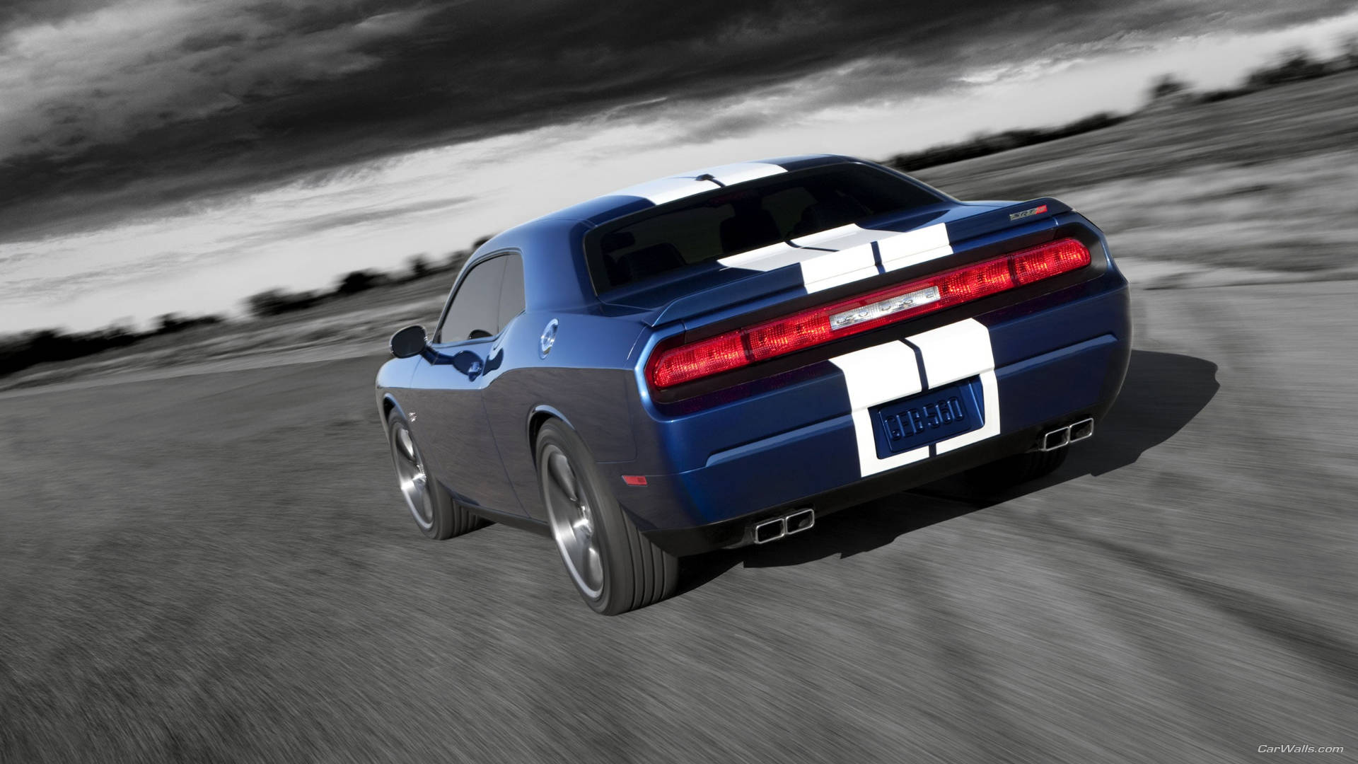 dodge challenger 桌布 壁紙 — 一輛帶有白色賽車條紋的藍色肌肉車在暴風雨的天空下，在模糊的沙漠公路上疾馳。