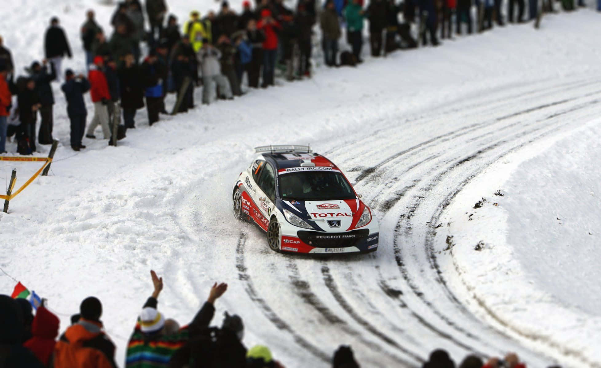 Ice Racing 桌布 壁紙 — 一輛帶有紅色和藍色貼紙的白色標緻拉力賽車在雪地彎道中高速行駛，留下深深的輪胎痕跡，觀眾在場邊歡呼。