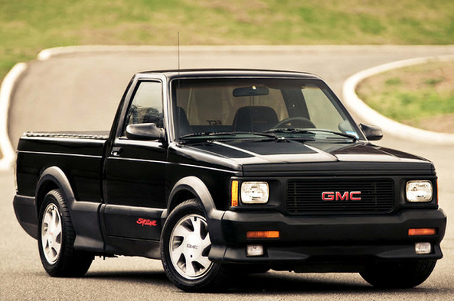Gmc Syclone 桌布 壁紙 — 一輛帶有紅色點綴的時尚黑色GMC Syclone皮卡車在彎曲的瀝青賽道上疾馳，賽道兩旁是草地。