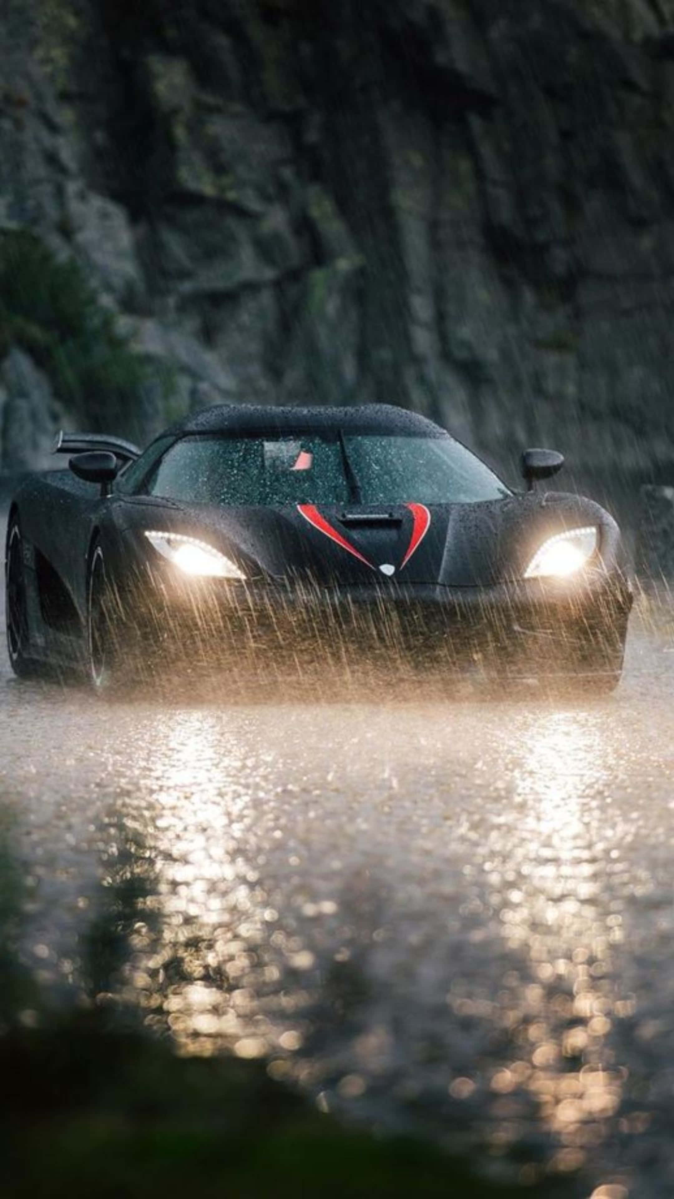 4K Car 桌布 壁紙 — 一輛帶有紅色點綴的時尚黑色跑車在岩石懸崖背景下飛馳過雨水濕滑的水面。