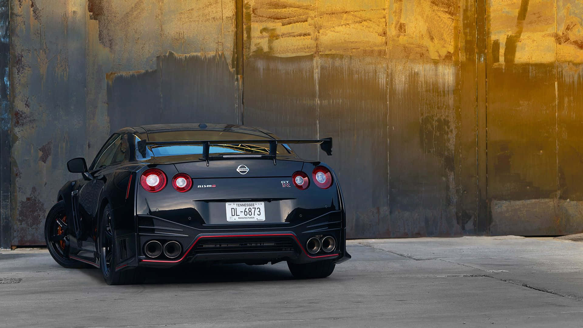 Gtr 桌布 壁紙 — 一輛帶有紅色點綴的黑色日產GT-R R35跑車，在金色陽光照耀下，停在一面風化的工業牆前。