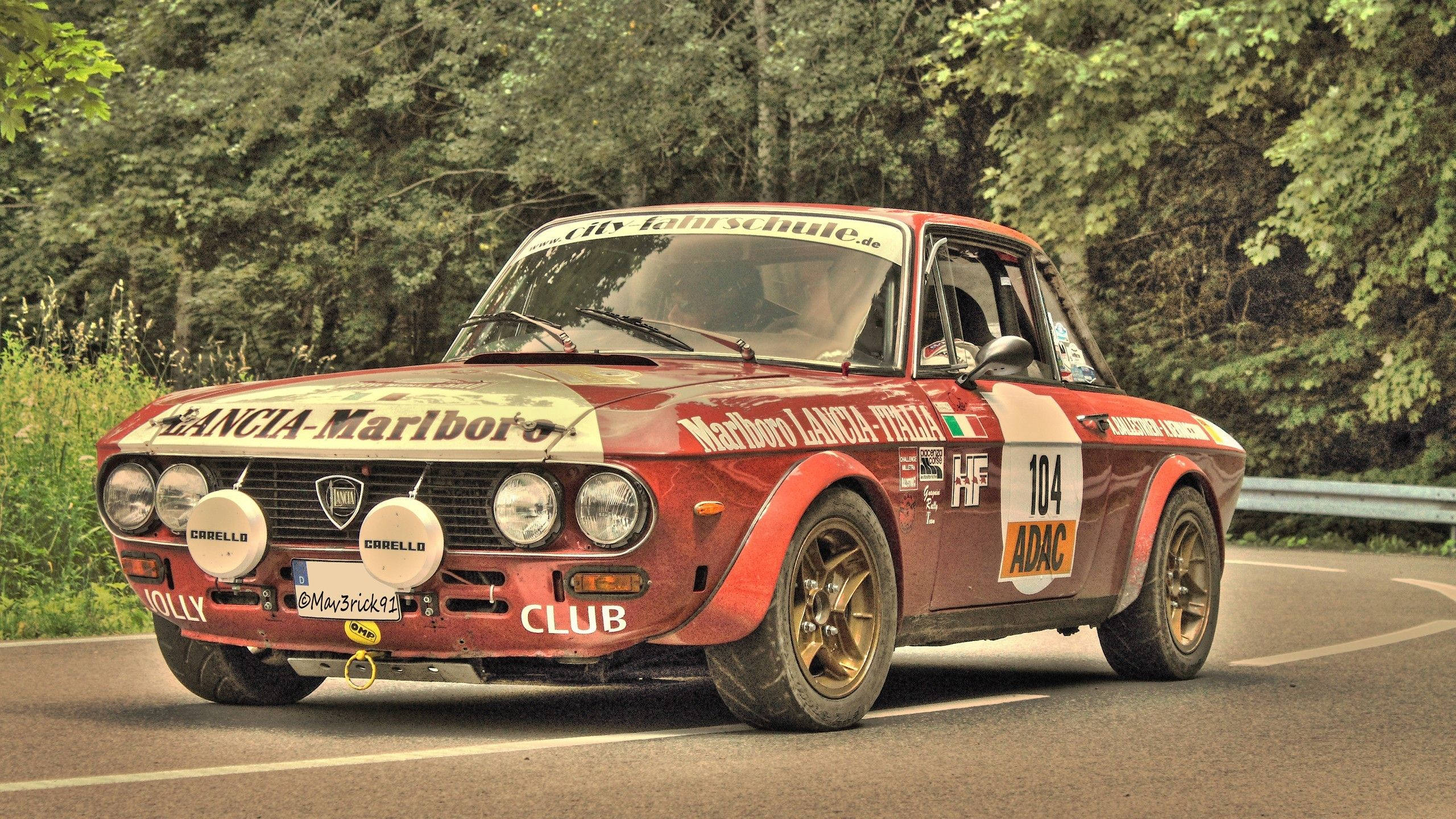 Red Lancia Fulvia Classic Car 壁紙 — 一輛帶有萬寶路貼紙的復古紅色藍旗亞拉力賽車在森林環繞的道路上飛馳。