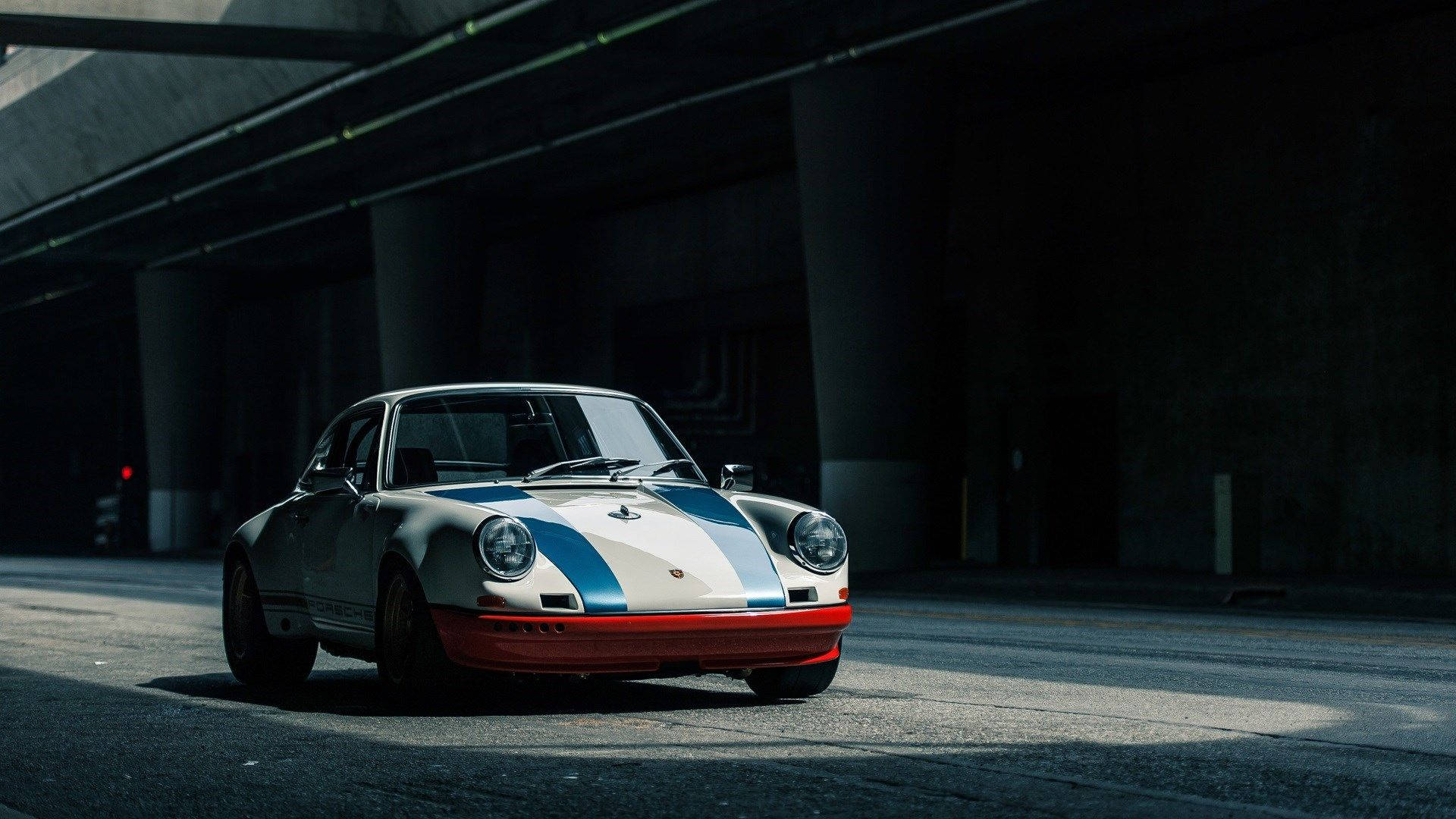 Classic Car White Porsche 911 壁紙 — 一輛帶有藍、白、紅條紋的復古保時捷911停在混凝土天橋下，沐浴在戲劇性的陽光中。