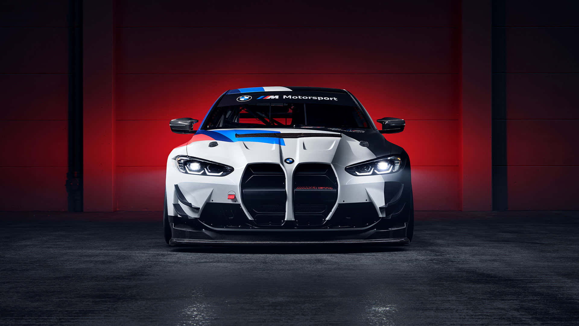 Sports Car 桌布 壁紙 — 一輛帶有藍色和紅色點綴的白色BMW M Motorsport賽車，在黑暗的車庫中，在戲劇性的紅色燈光下直視前方。