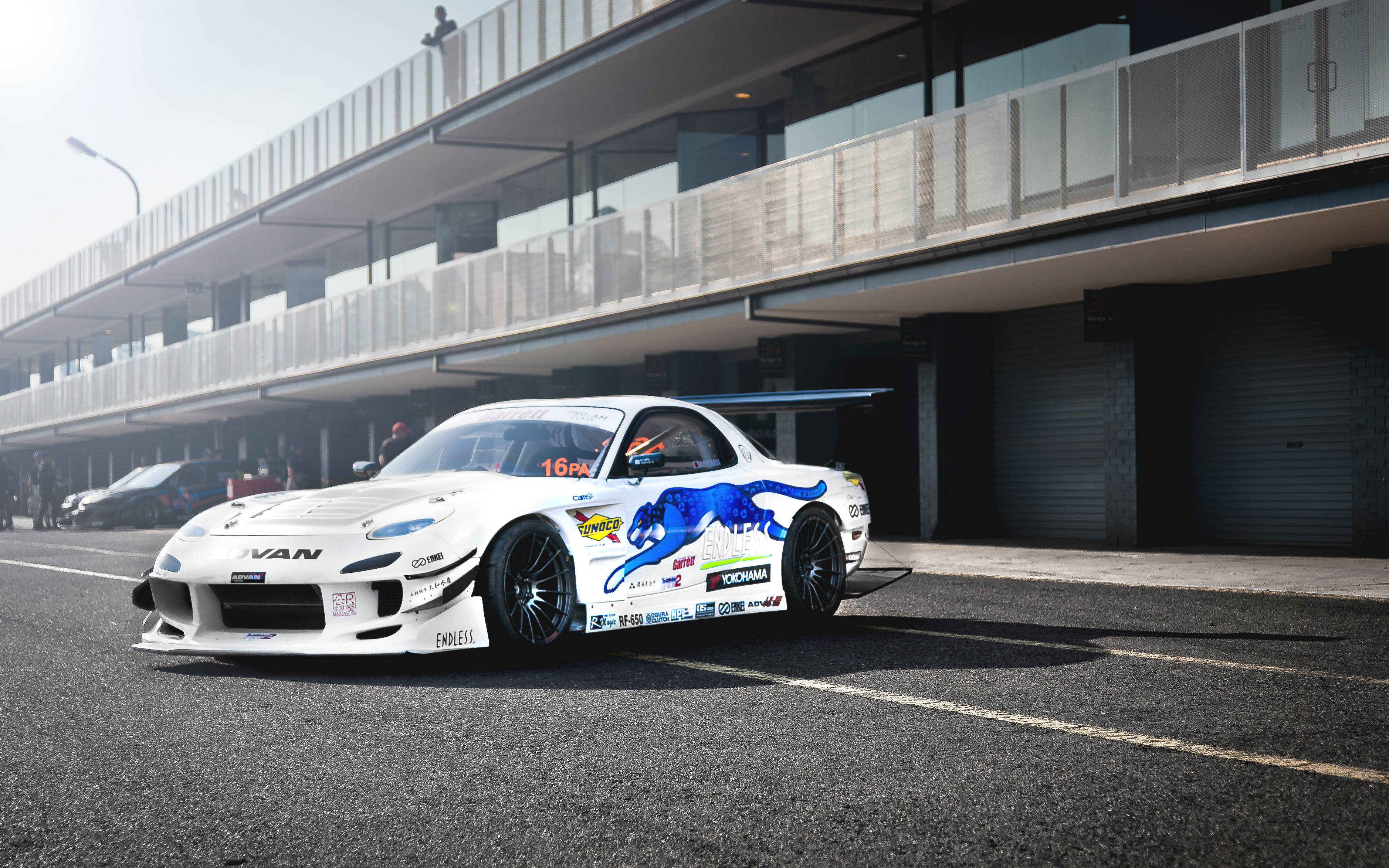 Mazda 桌布 壁紙 — 一輛帶有藍色恐龍圖案的白色漂移賽車停泊在賽車場的現代看台下。