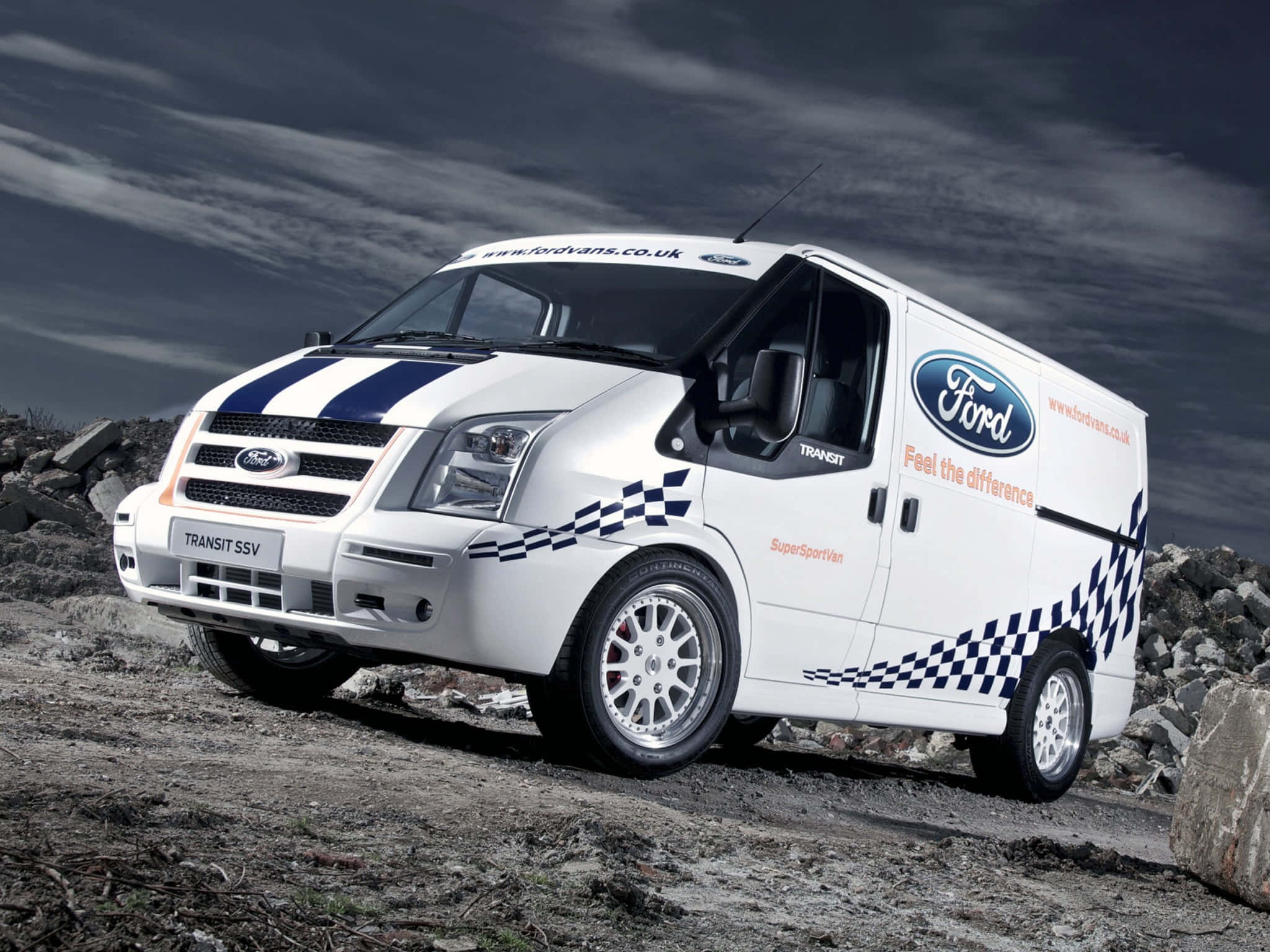 Ford Transit 桌布 壁紙 — 一輛帶有藍色賽車條紋和格子貼花的白色福特 Transit SSV 客貨車，站立在暴風雨天空下的岩石地形上。