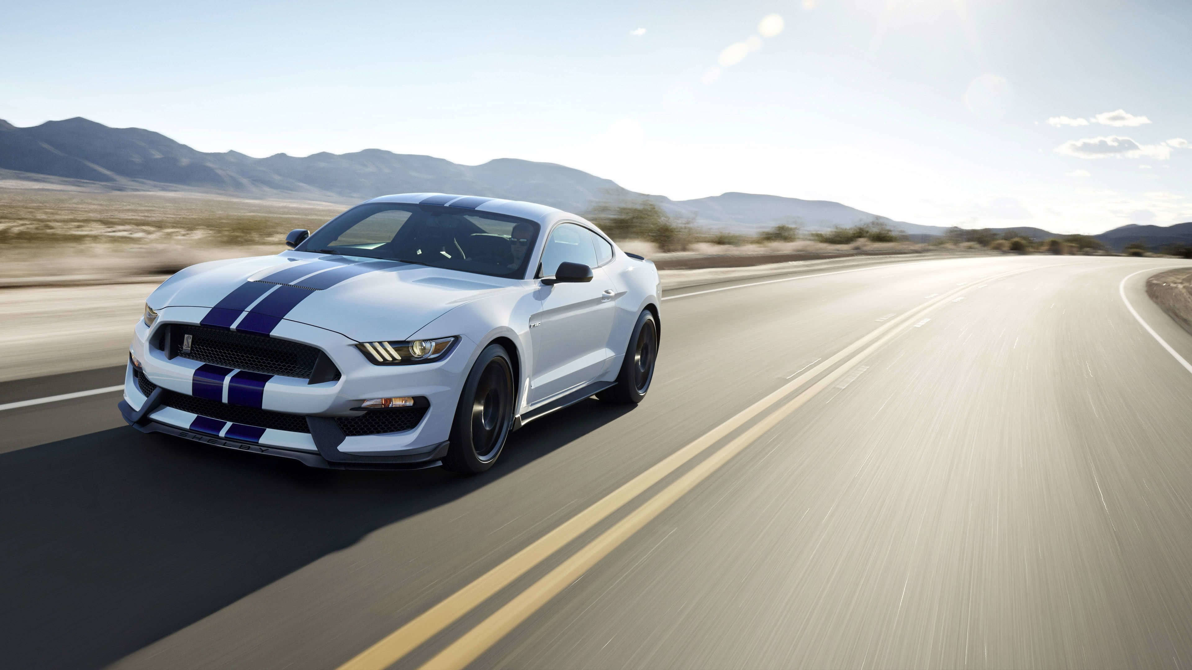mustang HD 桌布 壁紙 — 一輛帶有藍色賽車條紋的白色福特野馬在沙漠高速公路上疾馳，背景是山脈。