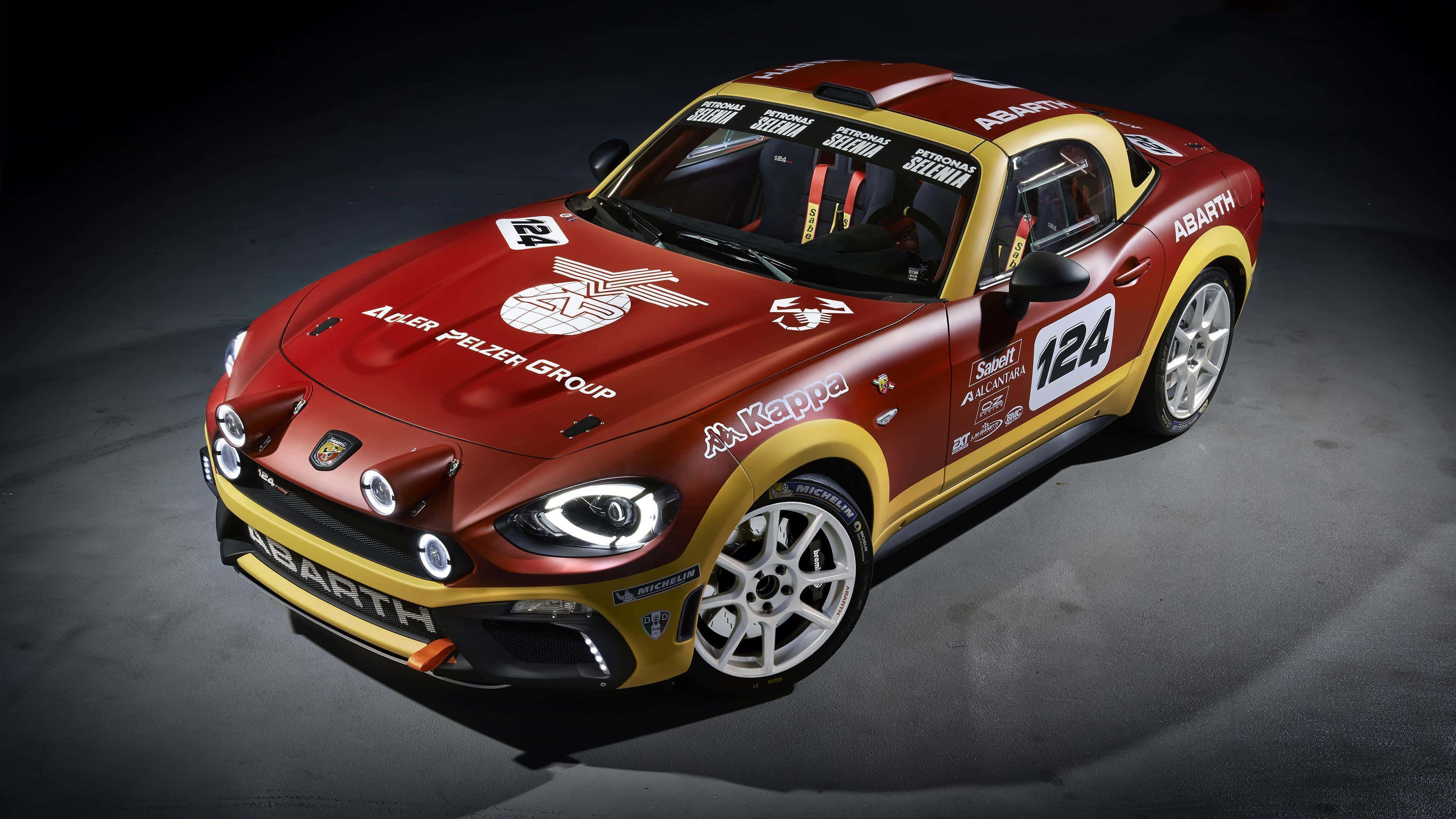 Abarth 桌布 壁紙 — 一輛帶有贊助商貼紙的紅色和金色 Abarth 124 賽車，在黑暗的攝影棚地板上被戲劇性地照亮。