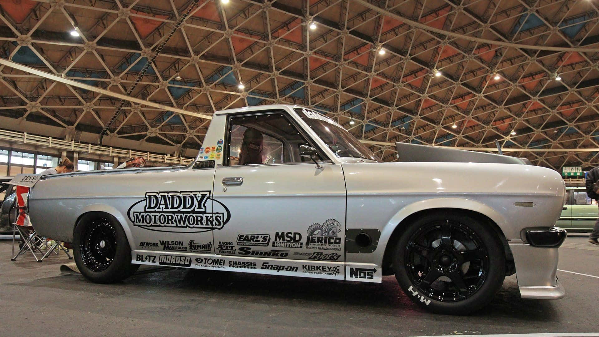 Datsun 桌布 壁紙 — 一輛帶有贊助商貼紙的銀色改裝皮卡車在幾何屋頂結構下室內展示。