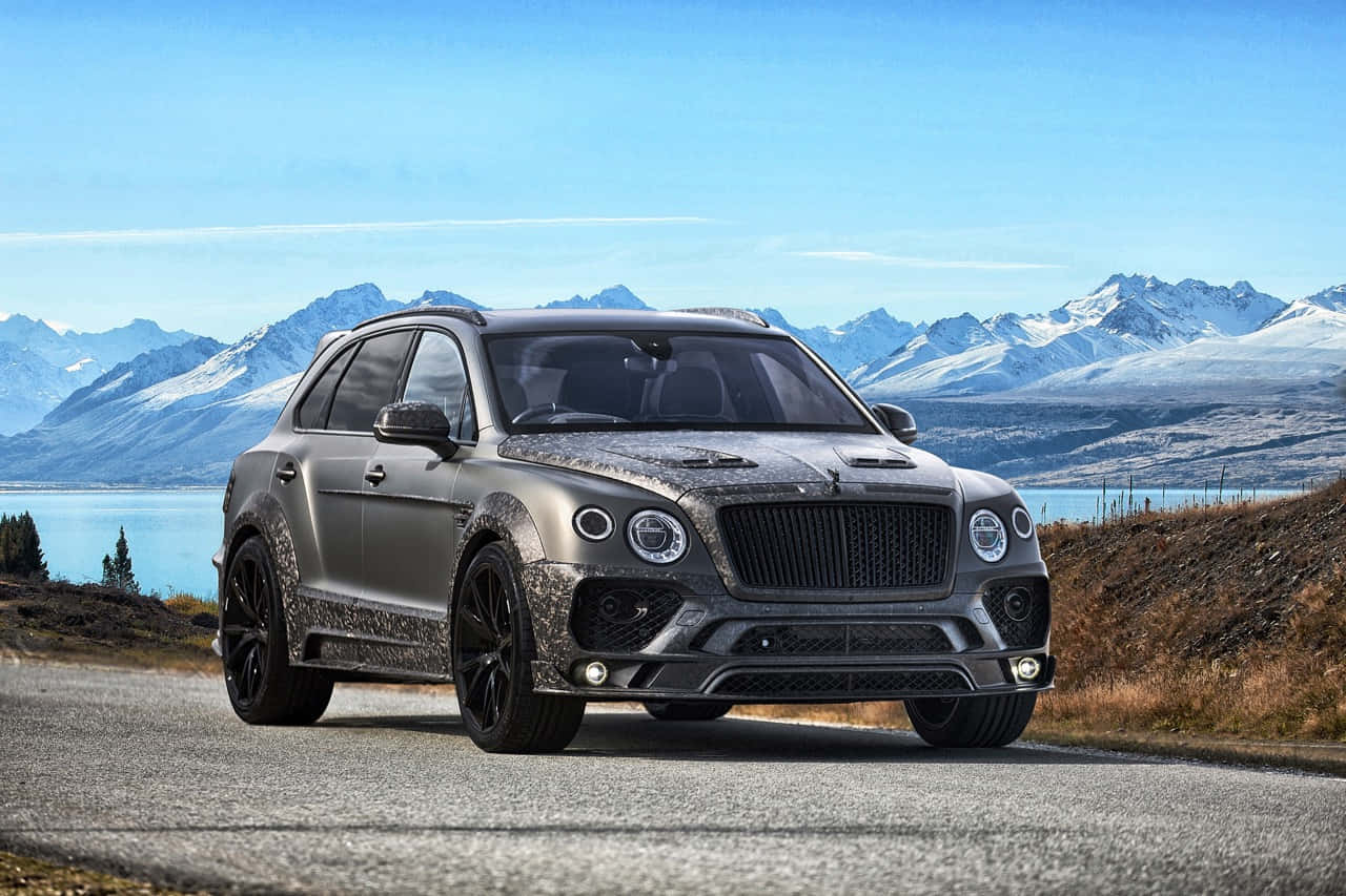 Bentley Bentayga 桌布 壁紙 — 一輛帶有迷彩細節的啞光灰色賓利 Bentayga SUV 在湖邊的鋪裝道路上行駛，背景是白雪覆蓋的山脈和清澈的藍天。