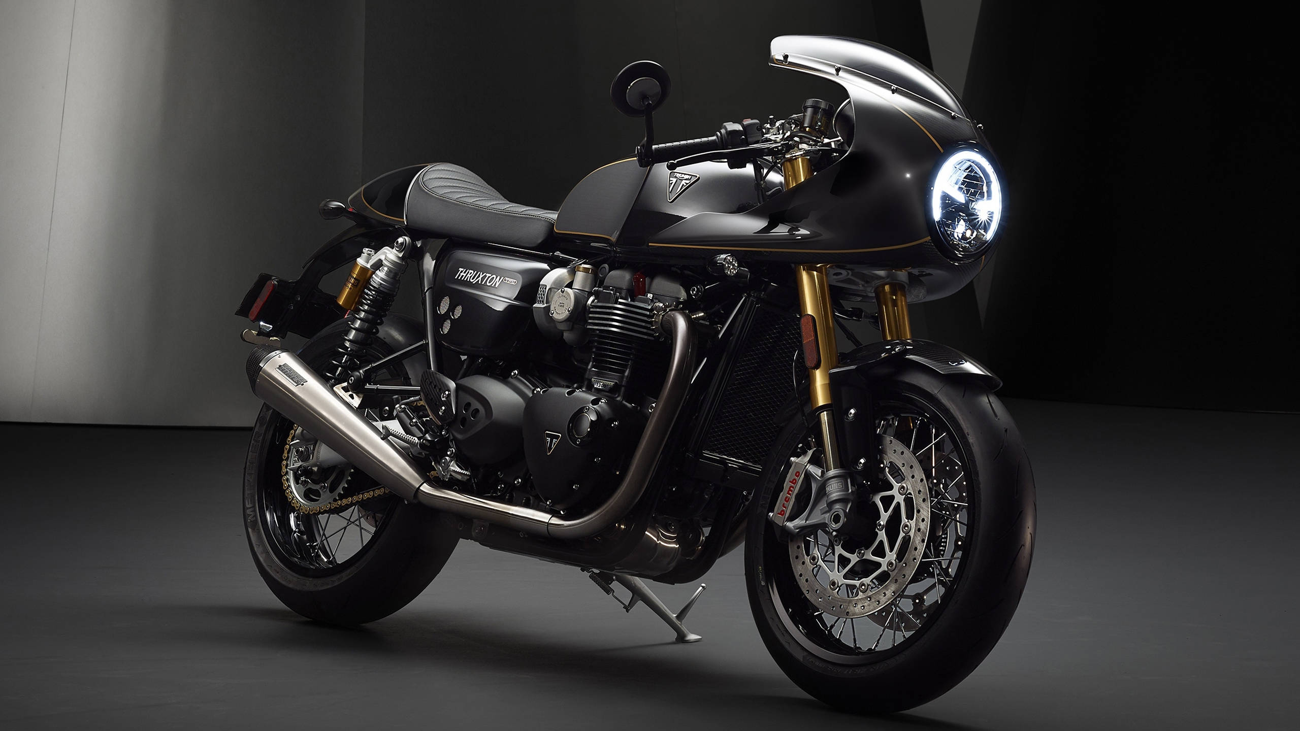 Triumph Bonneville Thurxton Jet Black 壁紙 — 一輛帶有金色點綴和發光車頭燈的時尚黑色 Triumph 電單車，在簡約的攝影棚環境中蓄勢待發。