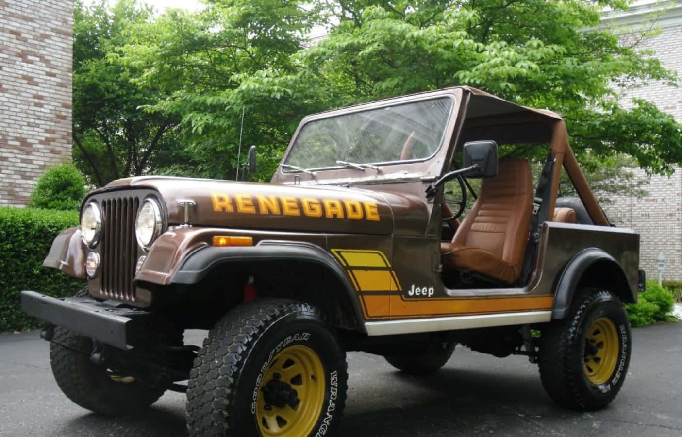 Jeep Cj 桌布 壁紙 — 一輛帶有黃色點綴和米色內飾的棕色Jeep Renegade停在瀝青路上，周圍環繞著綠樹和磚砌建築。