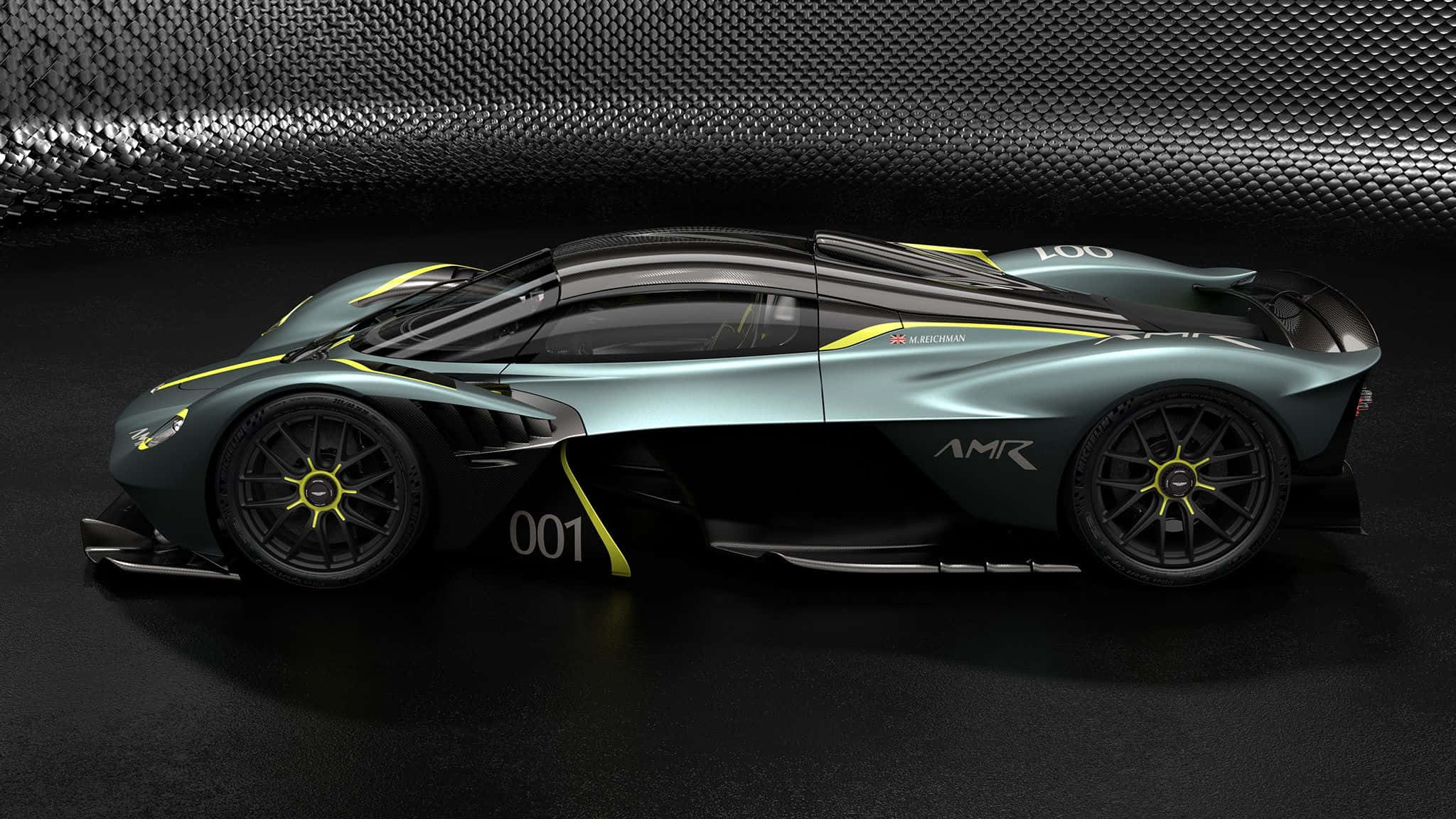 Aston Martin Valkyrie 桌布 壁紙 — 一輛帶有黃色點綴的時尚深綠色 Aston Martin AMR 賽車，在工作室環境中，以紋理金屬牆為背景展示。