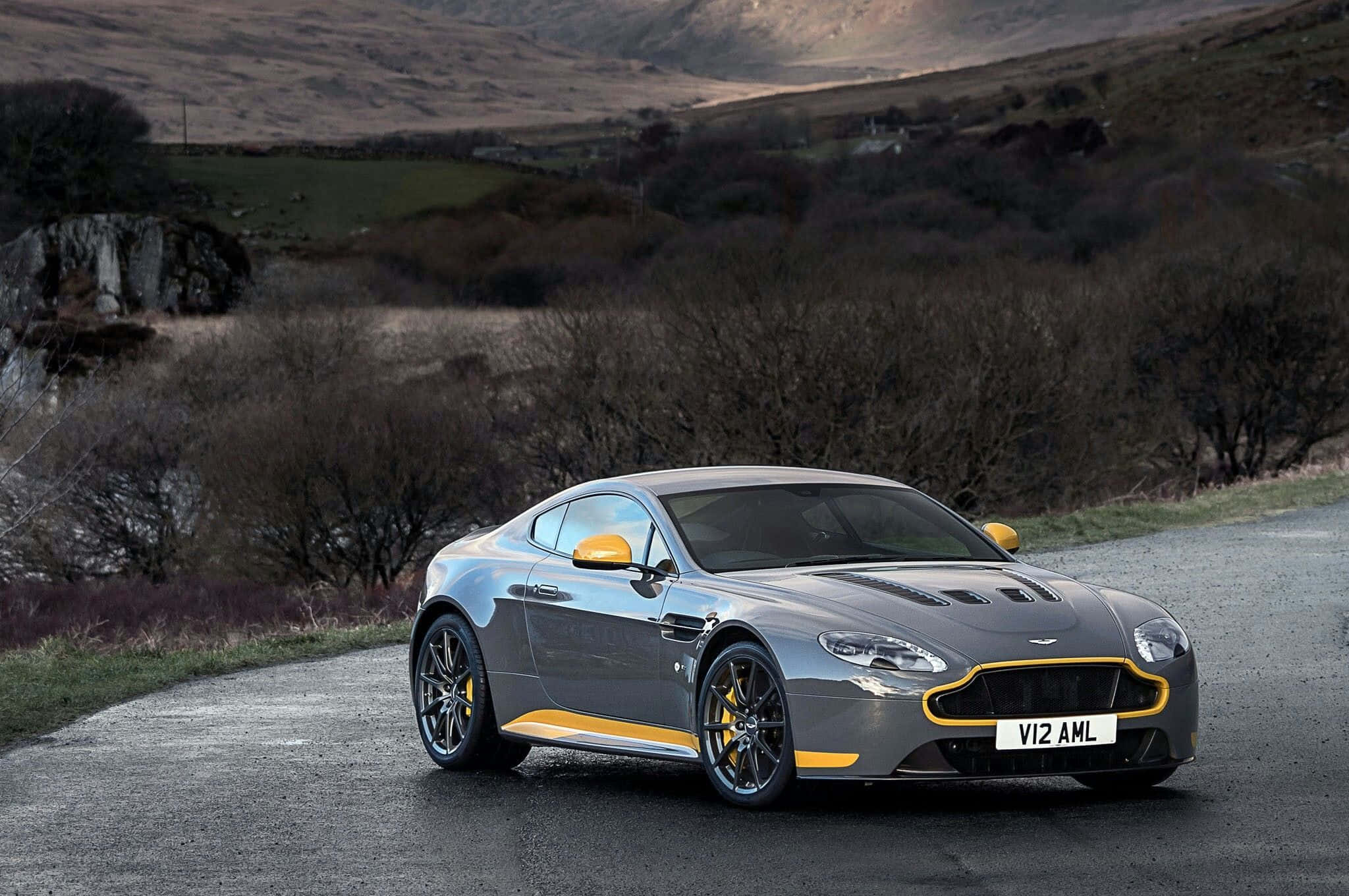 Aston Martin V12 Vantage 桌布 壁紙 — 一輛帶有黃色點綴的時尚銀色阿斯頓馬丁在蜿蜒的鄉村道路上穿過丘陵地帶。