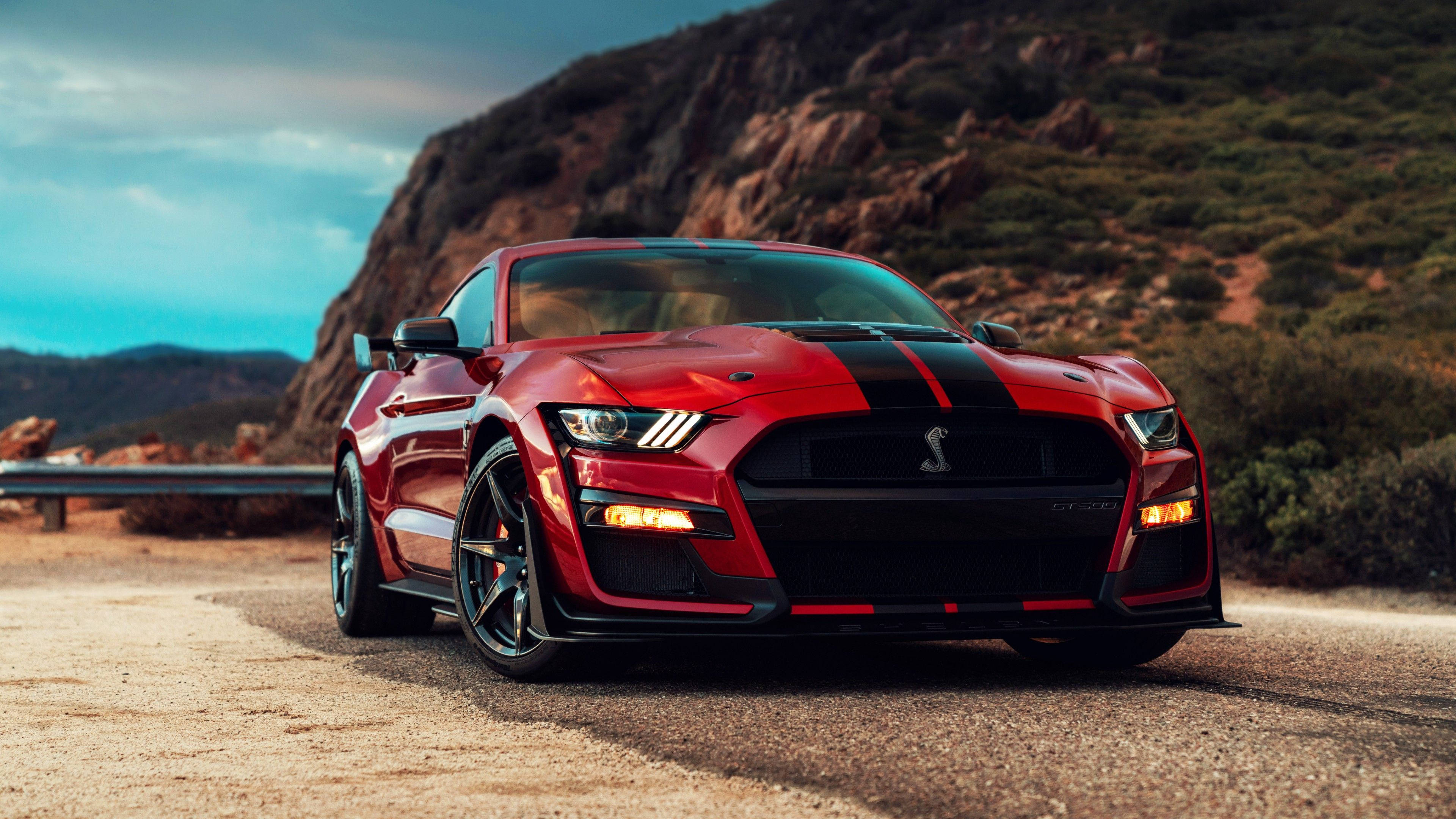 mustang HD 桌布 壁紙 — 一輛帶有黑色賽車條紋的紅色福特野馬謝爾比GT500停在蜿蜒的沙漠公路上，背景是岩石丘陵，天空多雲。