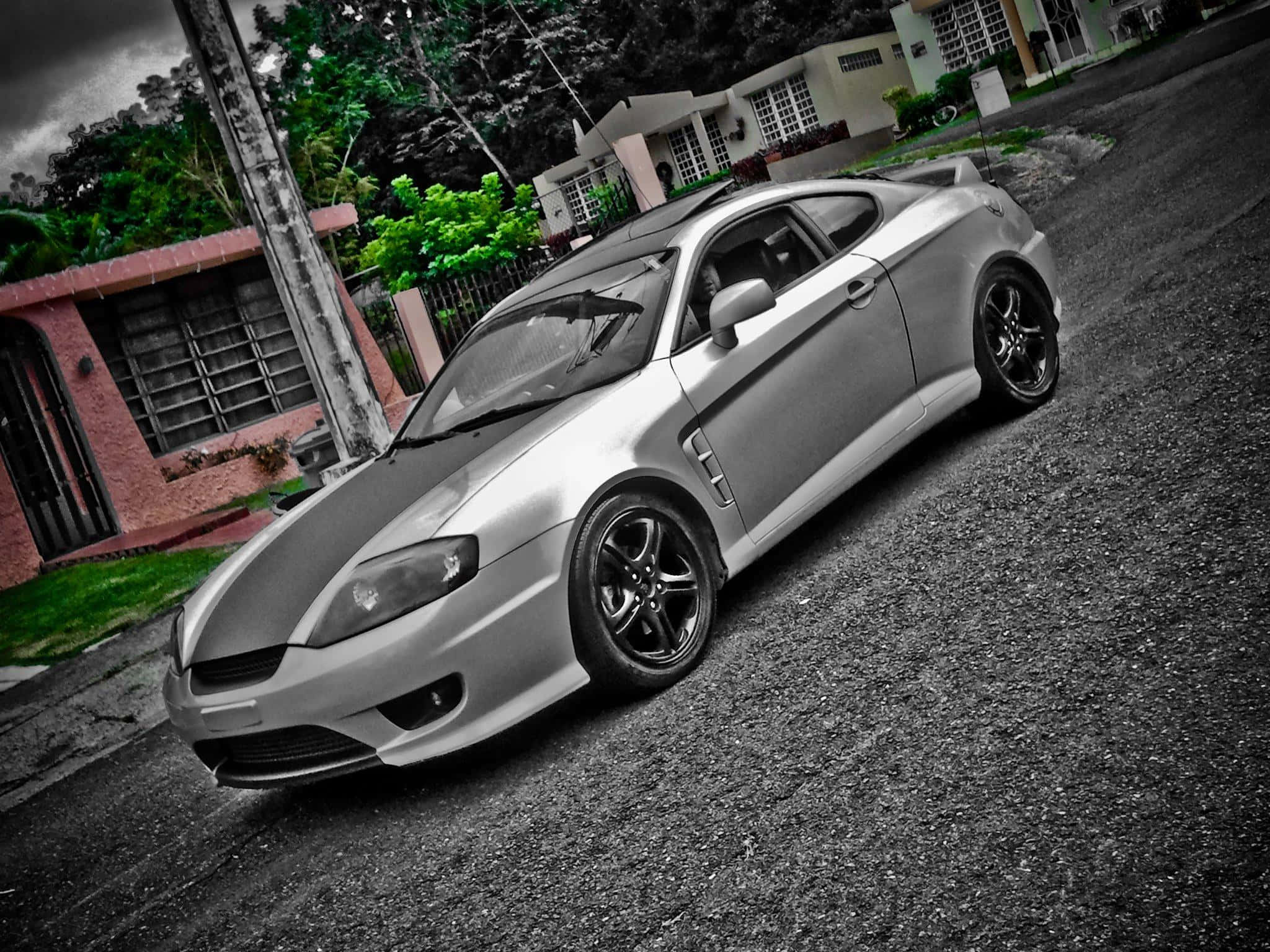 Hyundai Tiburon 桌布 壁紙 — 一輛帶黑色裝飾的銀色跑車停泊在碎石車道上，旁邊是一棟粉紅色的房子，天空多雲。