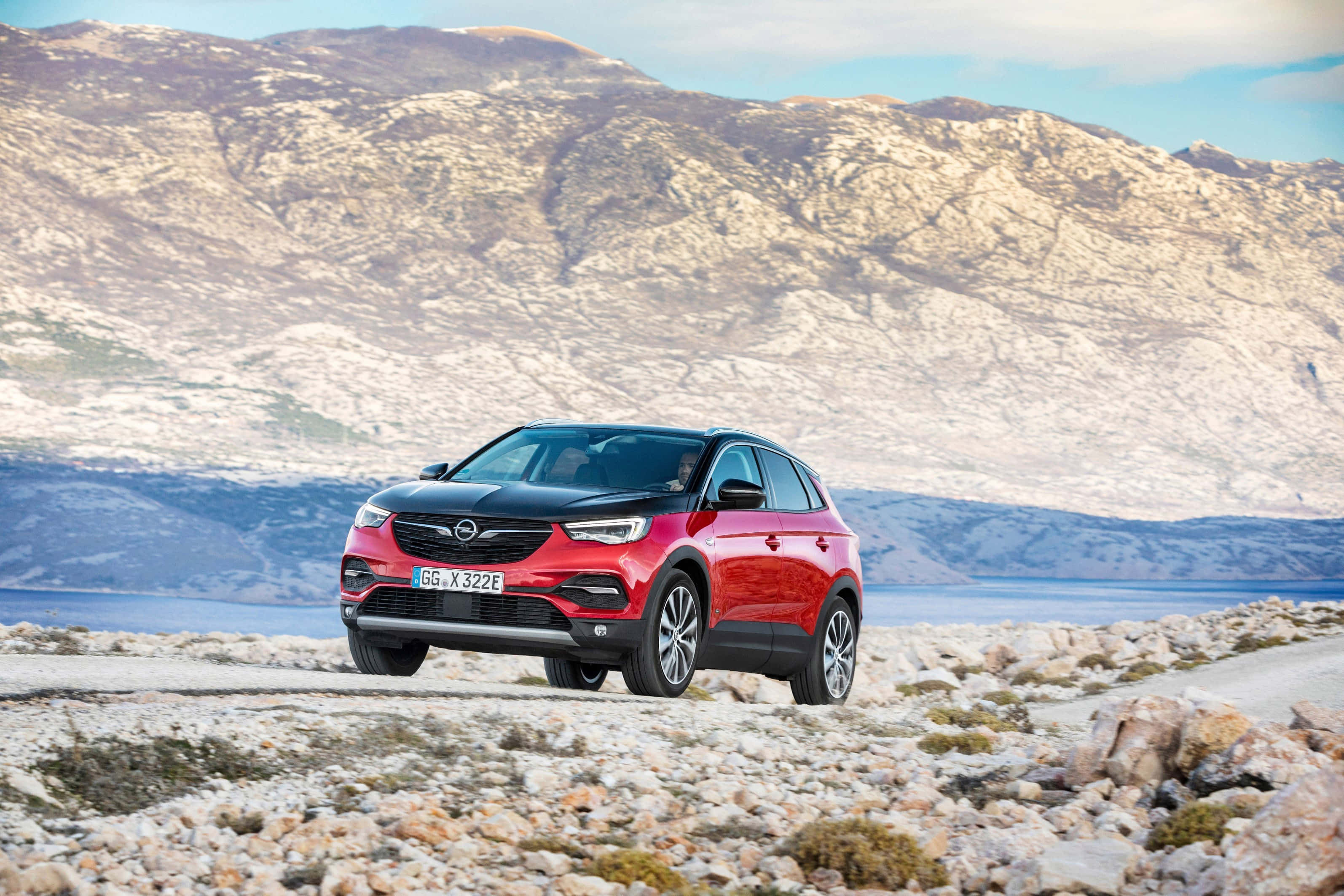 Opel Grandland X 桌布 壁紙 — 一輛引人注目的紅黑SUV停泊在岩石海岸道路上，背景是崎嶇的山脈和寧靜的大海。