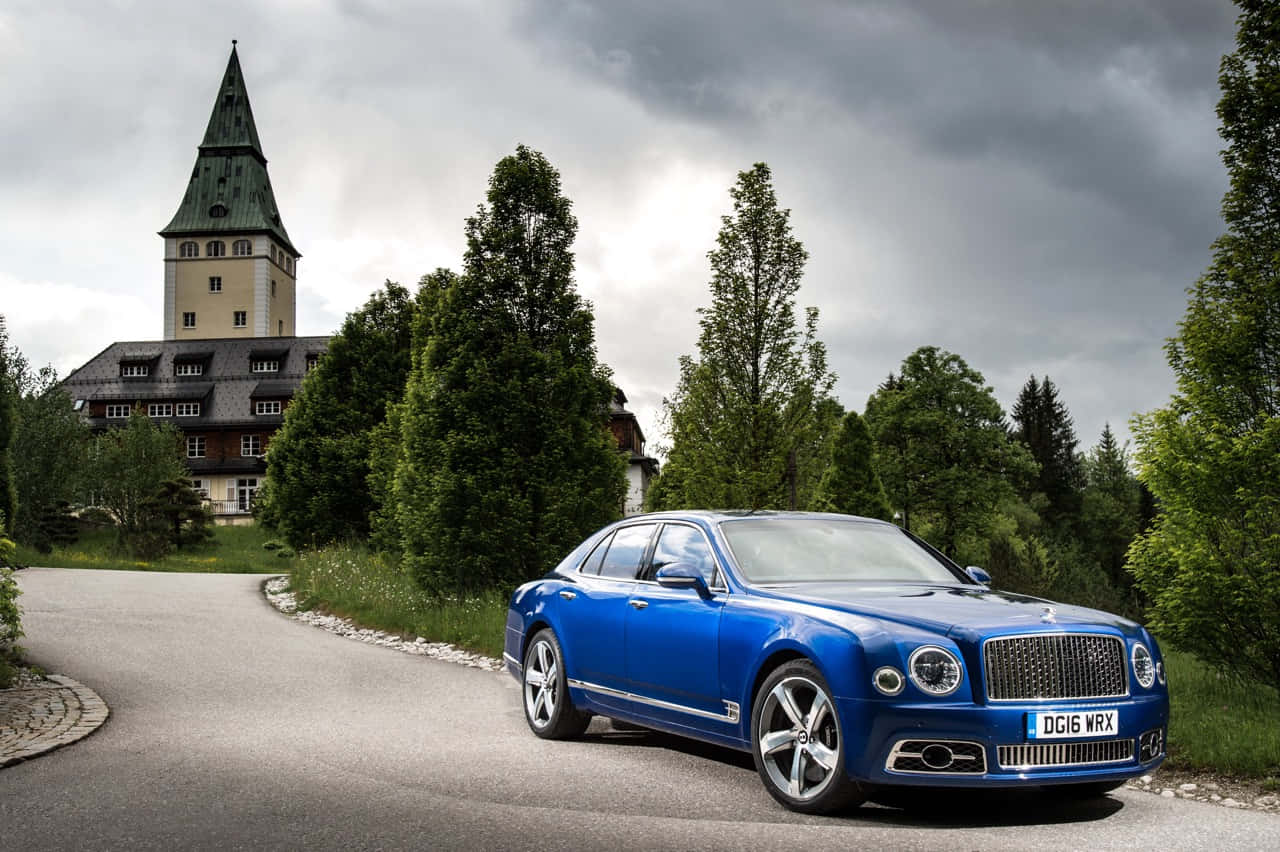 Bentley Mulsanne 桌布 壁紙 — 一輛引人注目的藍色賓利慕尚停泊在蜿蜒的道路上，前方是一座宏偉的歷史建築，天空陰沉多雲。
