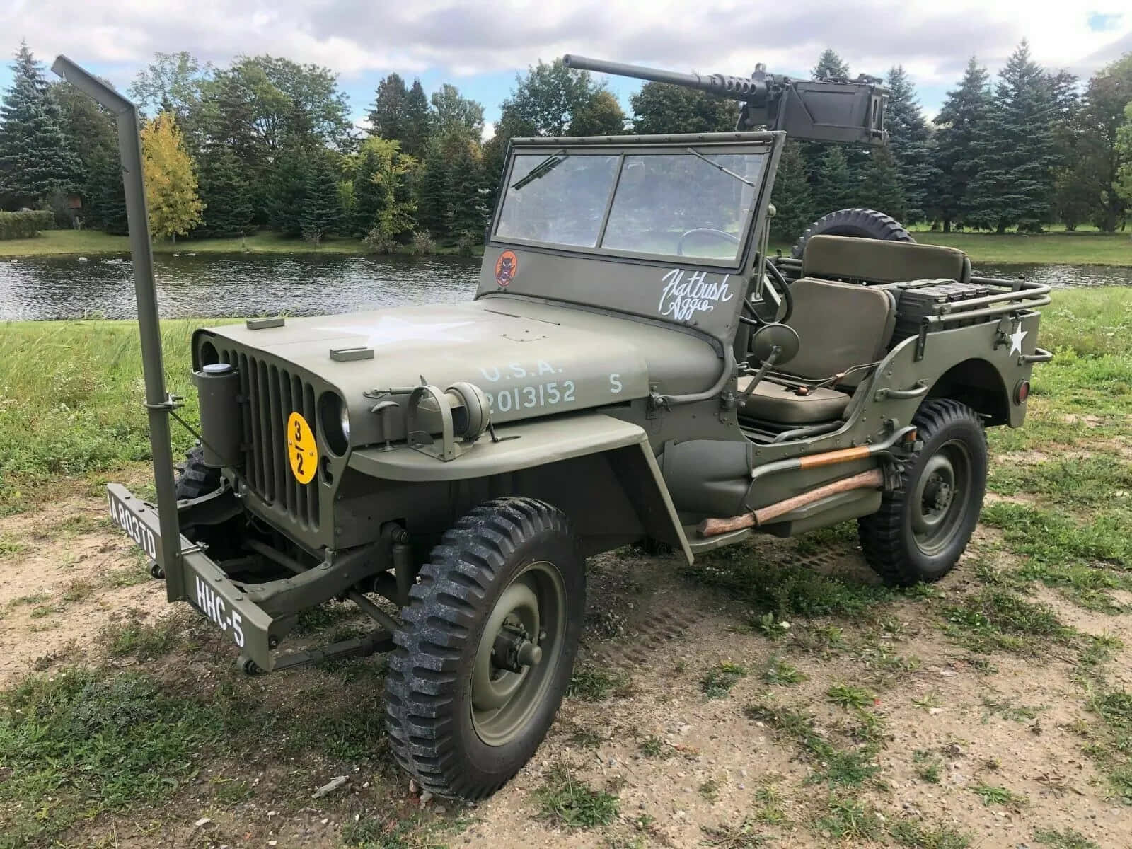 Jeep Willys 桌布 壁紙 — 一輛復古橄欖綠軍用吉普車，裝有機槍，停在樹木環繞的平靜湖泊附近的草地上。