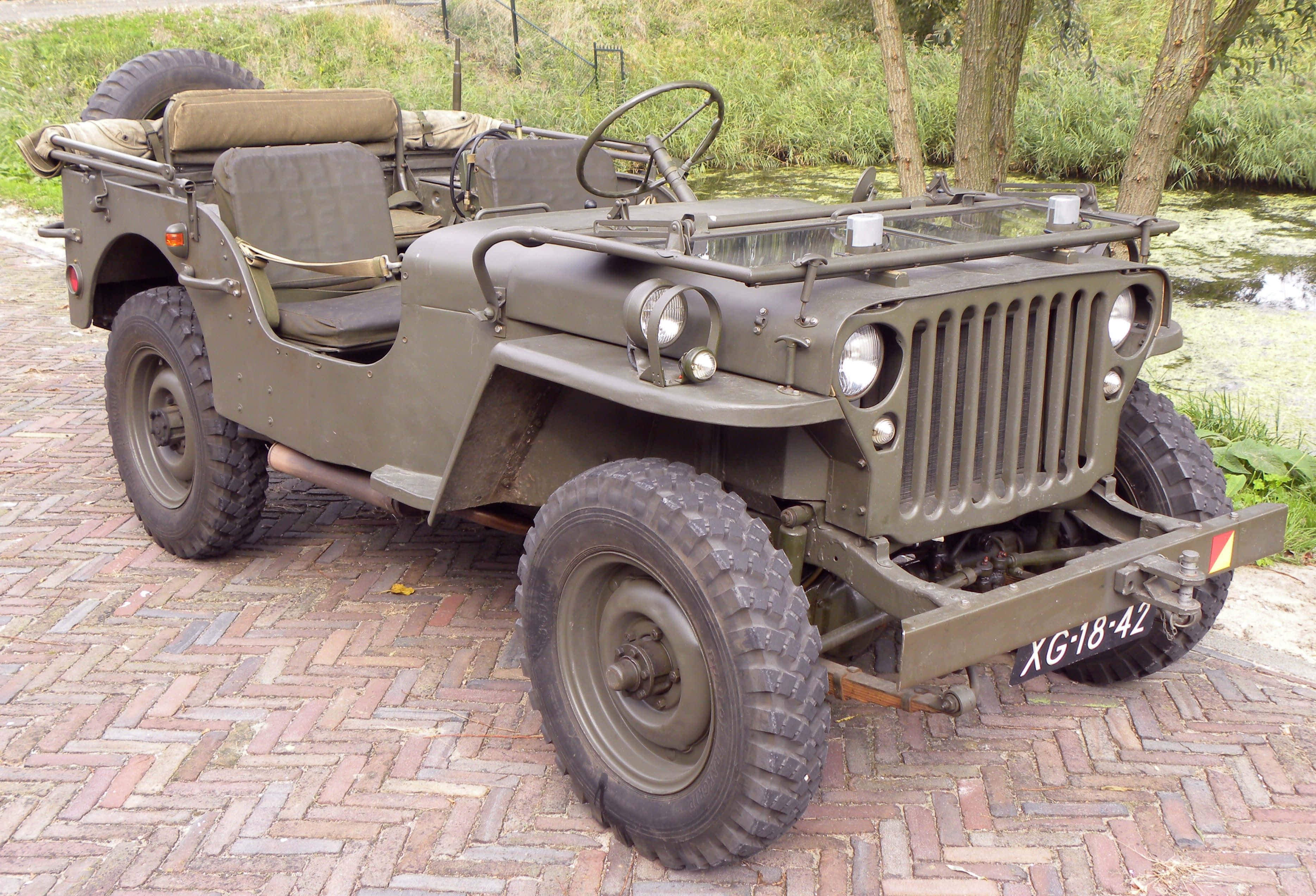 Jeep Willys 桌布 壁紙 — 一輛復古橄欖綠軍用吉普車停在紅磚小徑上，旁邊是帶有樹木和水的草地。