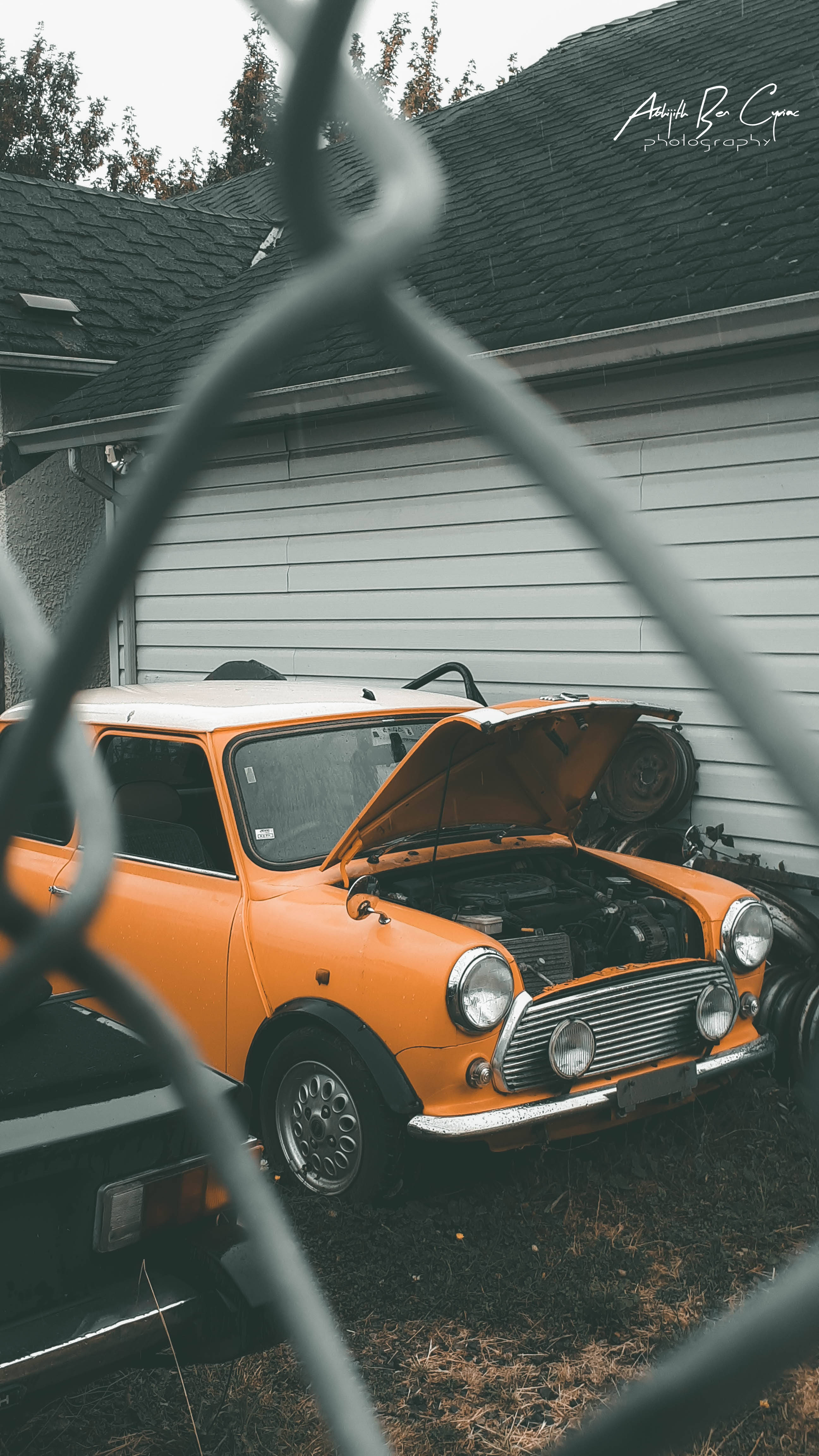 Vintage 90s Yellow Vehicle Aesthetic 壁紙 — 一輛復古橙色Mini Cooper打開引擎蓋，停在房子旁邊草地上的鐵絲網圍欄後面。