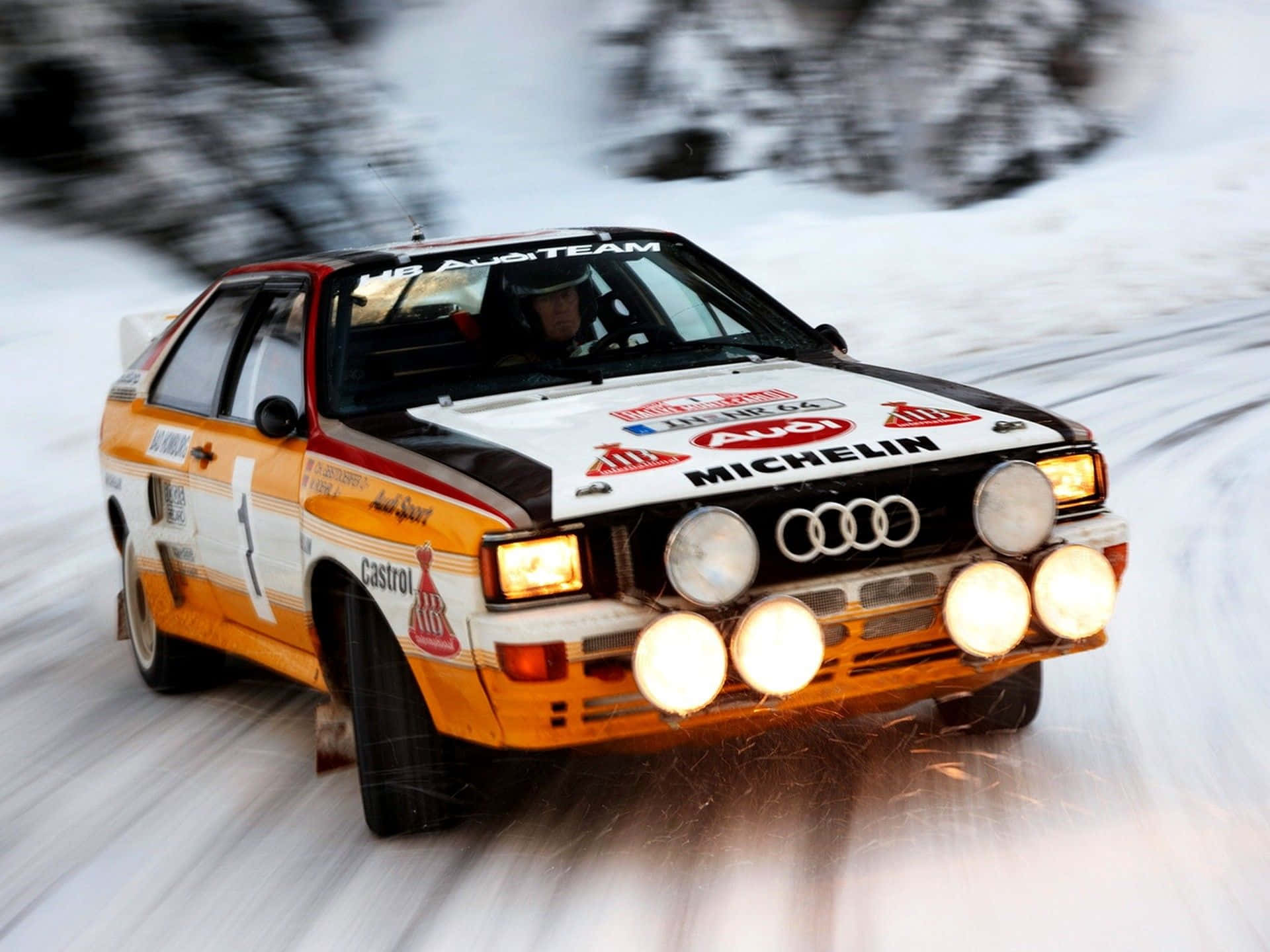 Ice Racing 桌布 壁紙 — 一輛復古橙色奧迪拉力賽車在雪地森林中疾馳，車頭燈亮起，身後雪花飛濺。