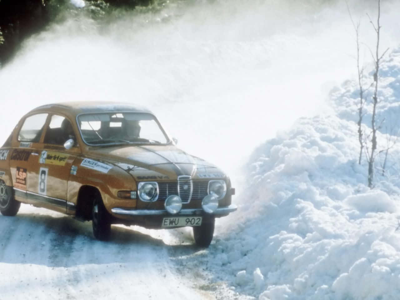 Ice Racing 桌布 壁紙 — 一輛復古橙色拉力賽車在雪地森林賽道上疾馳，揚起雪花。