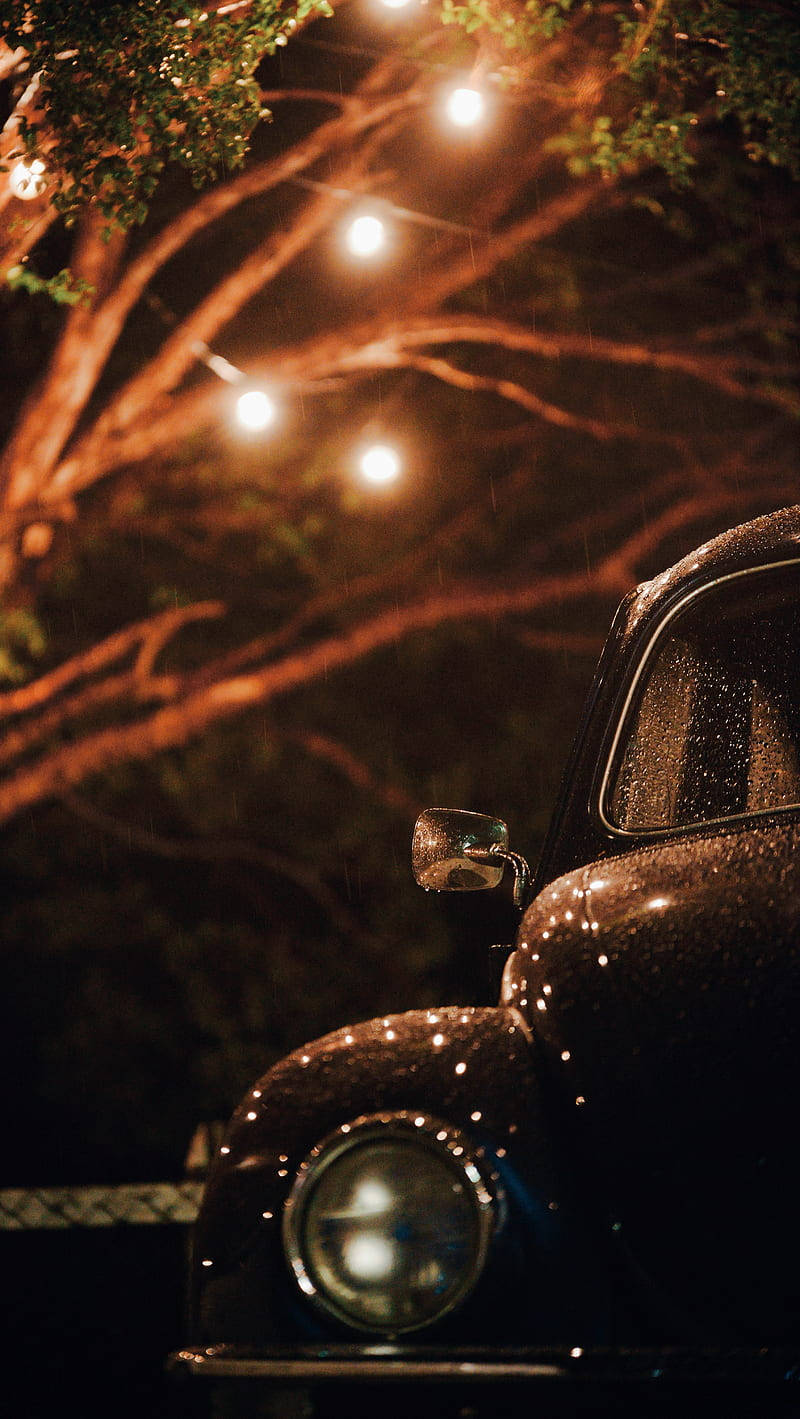 Retro 桌布 壁紙 — 一輛復古汽車在夜間的樹下閃爍著燈串的光芒，雨滴在引擎蓋上閃閃發光。