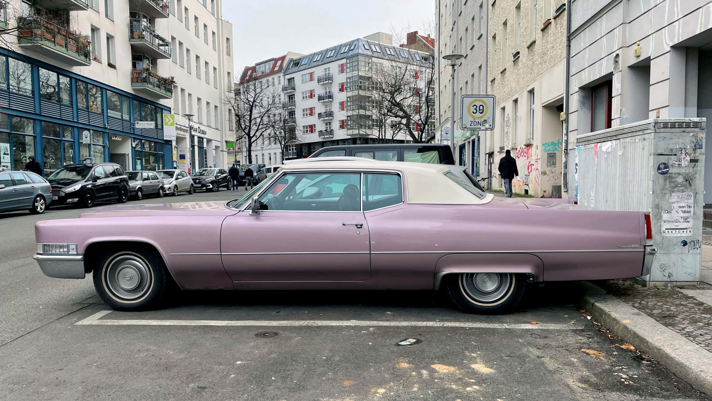 Pink Cadillac 桌布 壁紙 — 一輛復古淡紫色凱迪拉克，白色車頂，停泊在現代建築和光禿樹木林立的城市街道上。