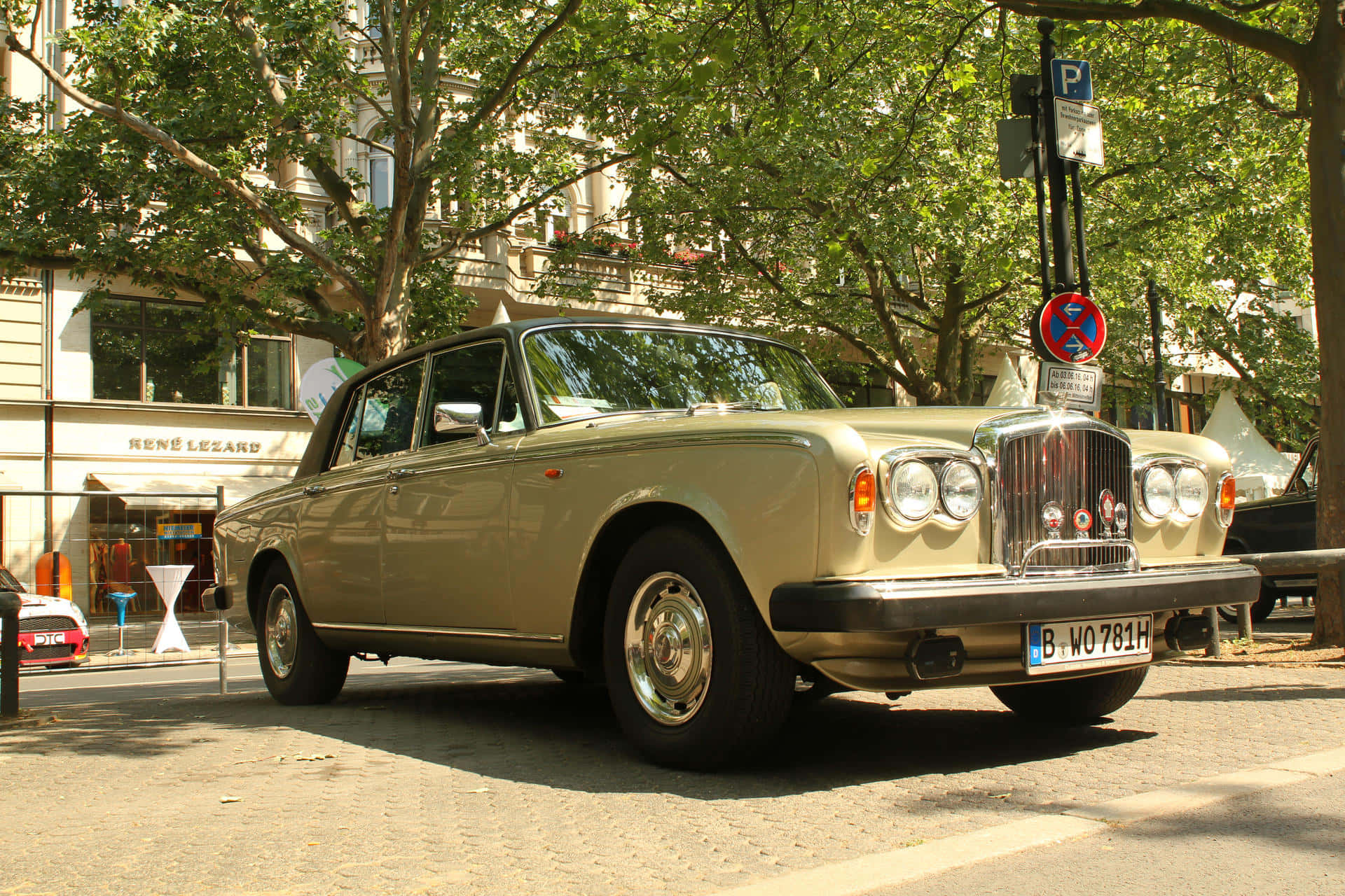 Bentley T2 桌布 壁紙 — 一輛復古米色賓利轎車停放在陽光普照、綠樹成蔭的城市街道上。