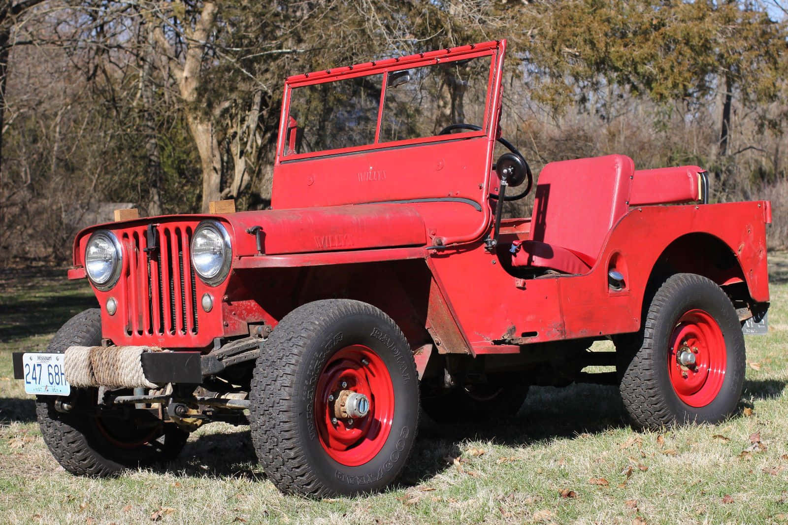 Jeep Willys 桌布 壁紙 — 一輛復古紅色軍用吉普車，配備堅固輪胎，停泊在陽光普照的田野中、光禿禿的樹木下的草地上。