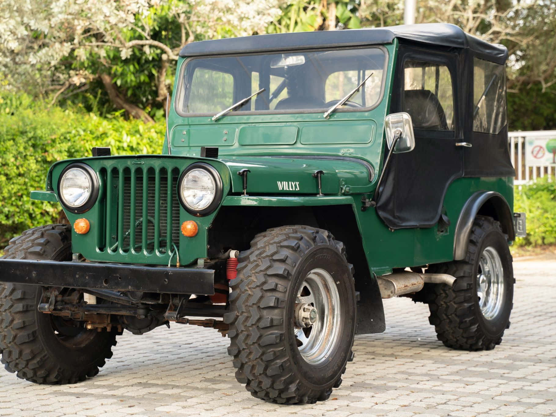 Jeep Willys 桌布 壁紙 — 一輛復古綠色Willys Jeep，配備黑色軟頂和堅固輪胎，停泊在鋪砌的庭院中，四周環繞著茂密的綠植。