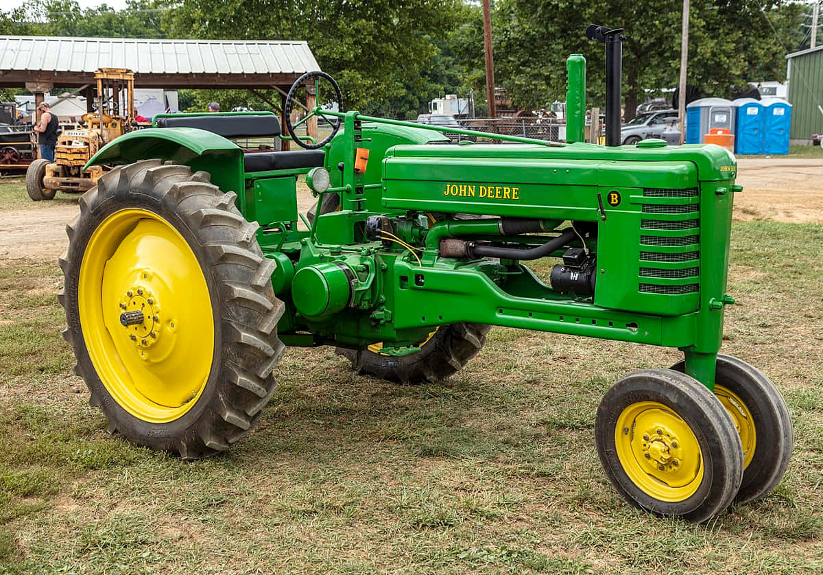 John Deere 桌布 壁紙 — 一輛復古綠色約翰迪爾B型拖拉機，配亮黃色車輪，自豪地矗立在戶外農機展的草地上。