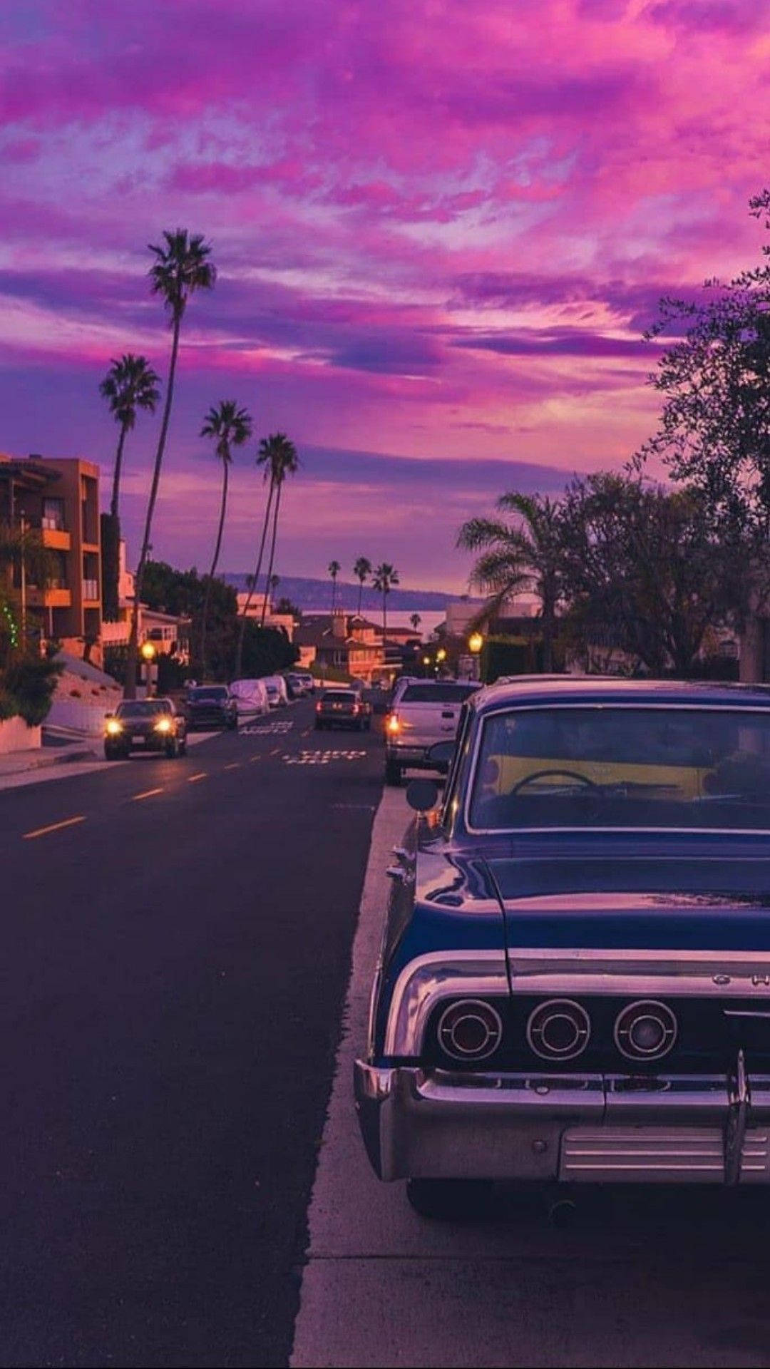 Pastel Phone Pastel Skies 壁紙 — 一輛復古藍色汽車停泊在棕櫚樹成蔭的街道上，背景是充滿活力的紫色和粉紅色日落天空。