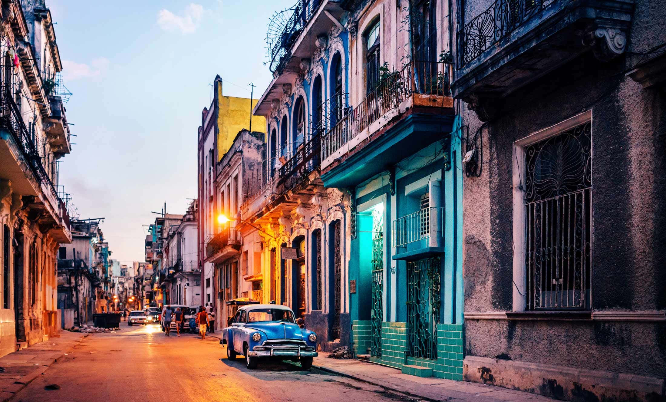 Cuba 桌布 壁紙 — 一輛復古藍色汽車在黃昏時分，在狹窄的古巴街道上，溫暖的路燈下閃耀，街道兩旁是色彩繽紛的殖民時期建築。