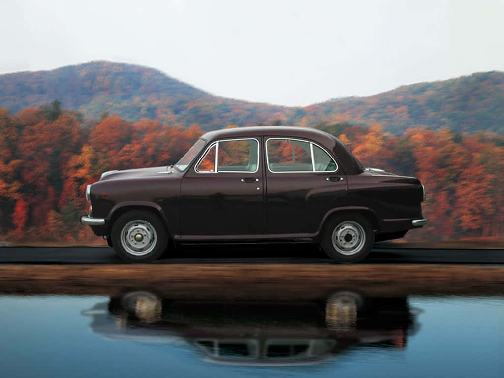 Hindustan Motors 桌布 壁紙 — 一輛復古酒紅色汽車在平靜的水面上完美倒映，背景是秋天的森林和起伏的山丘。