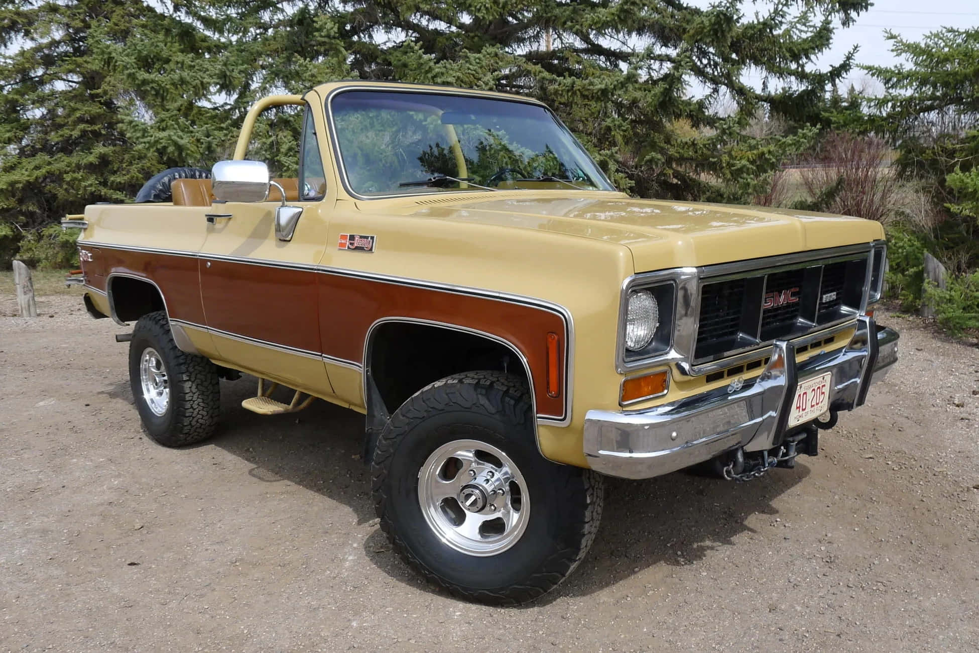 Gmc Jimmy 桌布 壁紙 — 一輛復古雙色黃色和棕色的GMC皮卡車，帶有鍍鉻裝飾和大輪胎，停在常綠樹附近的泥土上。