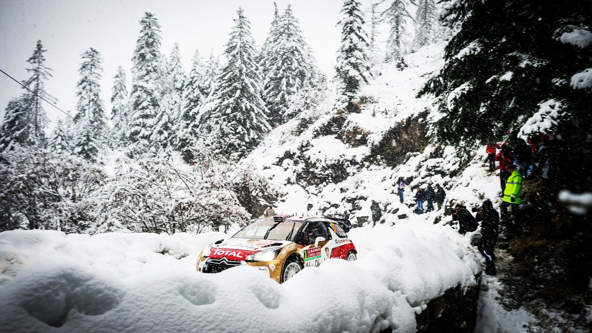 Ice Racing 桌布 壁紙 — 一輛拉力賽車在森林覆蓋的山路上與深雪搏鬥，觀眾從雪堆旁觀看。