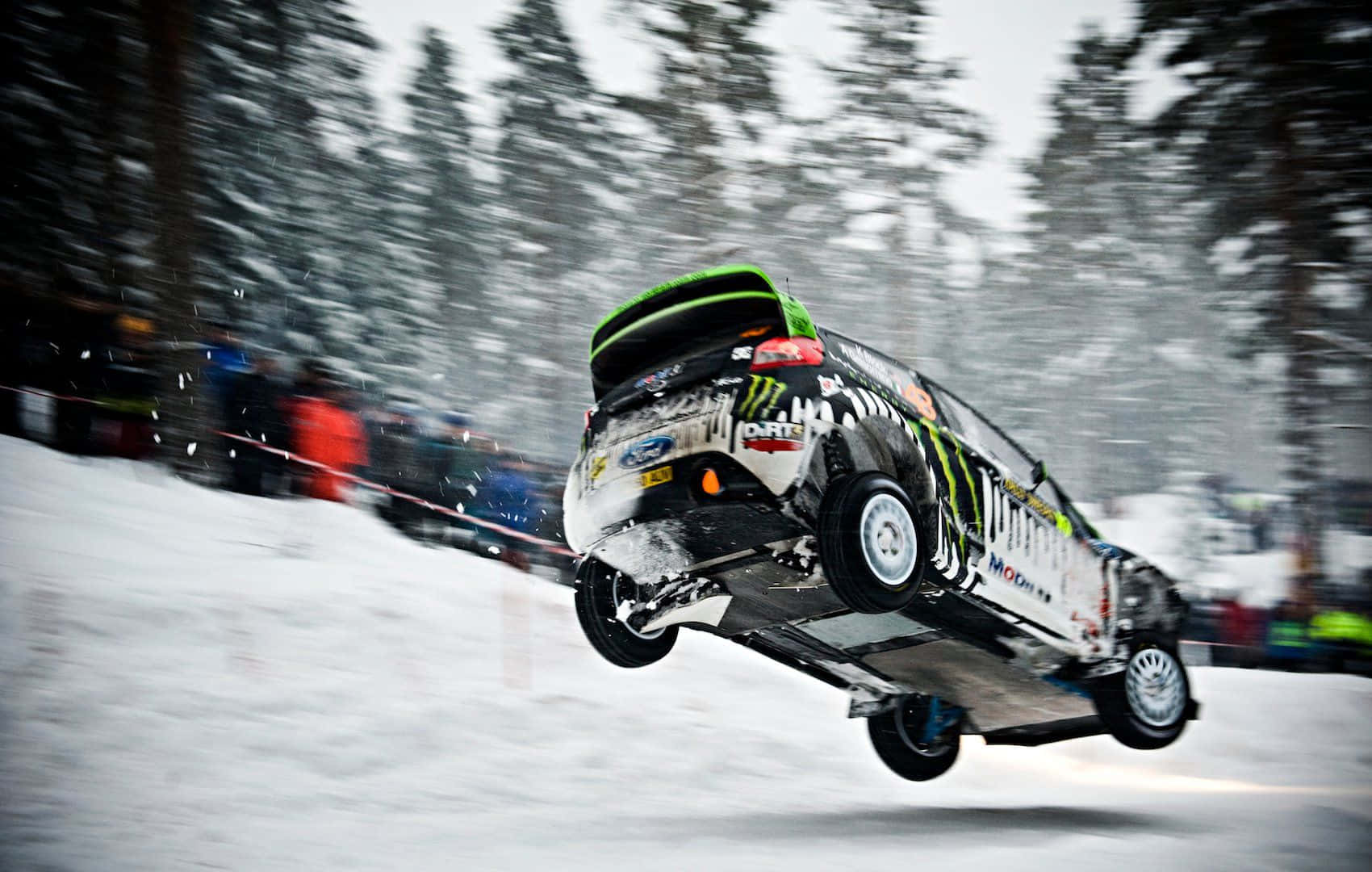 Ice Racing 桌布 壁紙 — 一輛拉力賽車在空中飛越雪地地形，松樹和觀眾因運動而模糊。