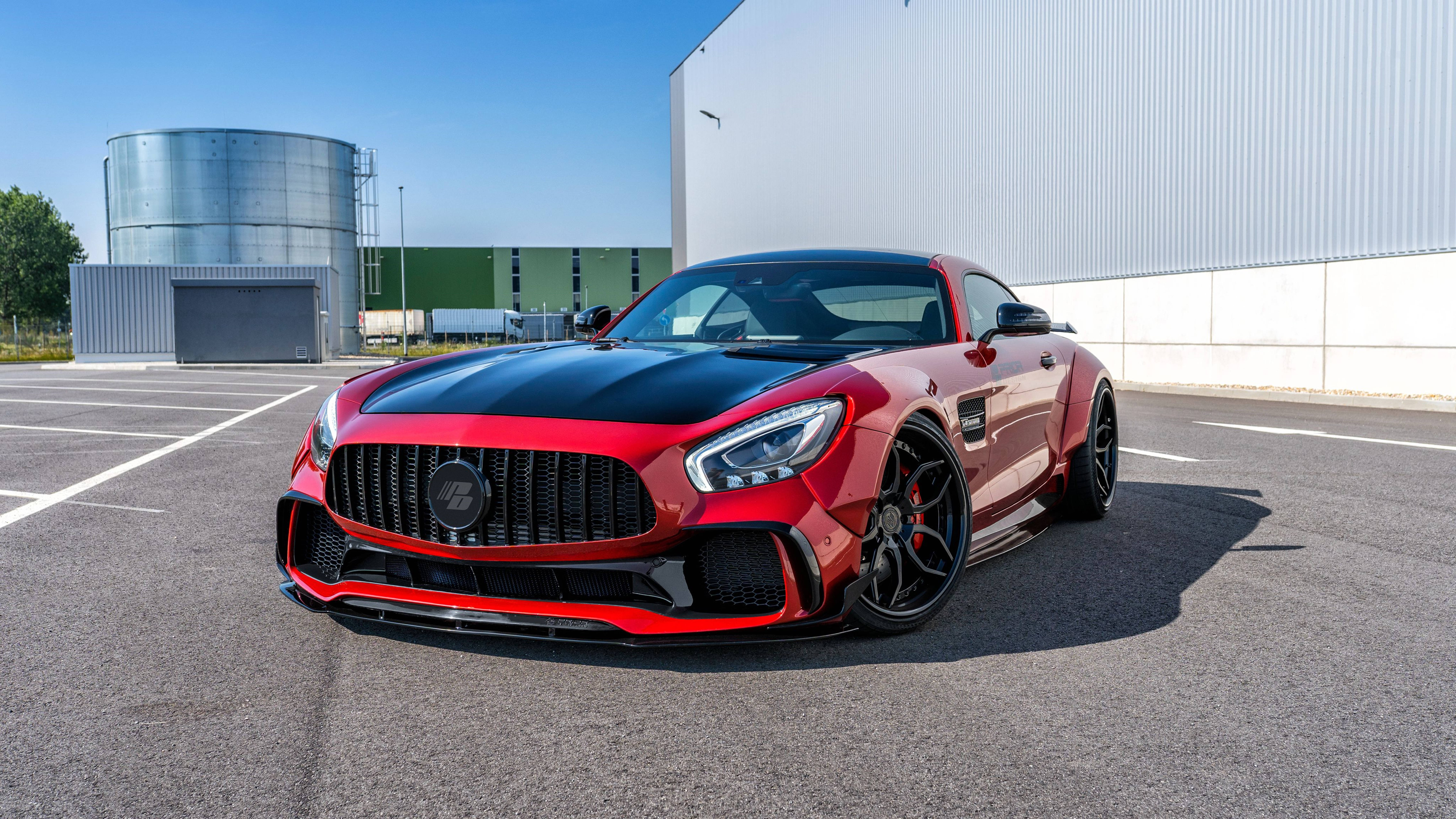 HD mercedes 桌布 壁紙 — 一輛搶眼的紅黑色 Mercedes-AMG GT R 跑車停泊在陽光普照的工業用地，背景有一個大型銀色儲罐和白色建築物。
