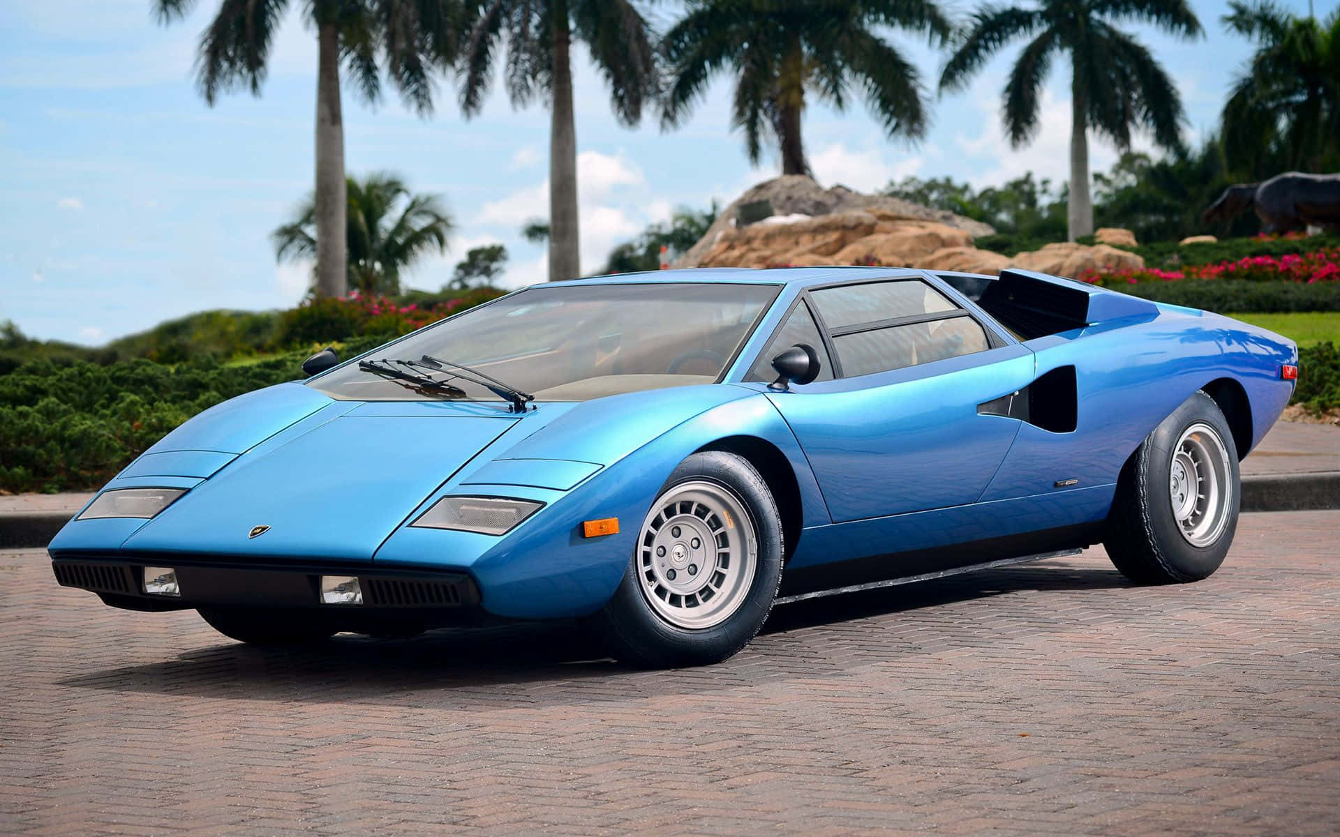 Lamborghini Countach 桌布 壁紙 — 一輛搶眼的藍色林寶堅尼Countach停泊在磚砌車道上，周圍有棕櫚樹和園林花園，天空晴朗。