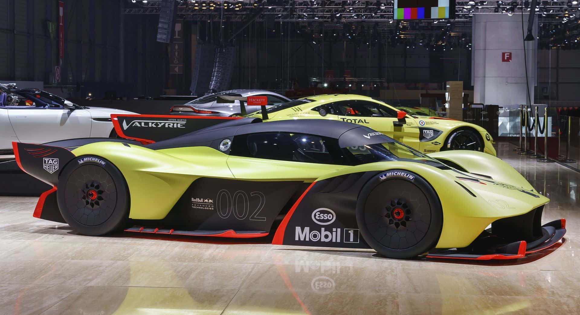 Aston Martin Valkyrie 桌布 壁紙 — 一輛搶眼的青檸綠色Aston Martin Valkyrie賽車，帶有黑色和紅色點綴，在車展上展出，背景是其他高性能車輛。